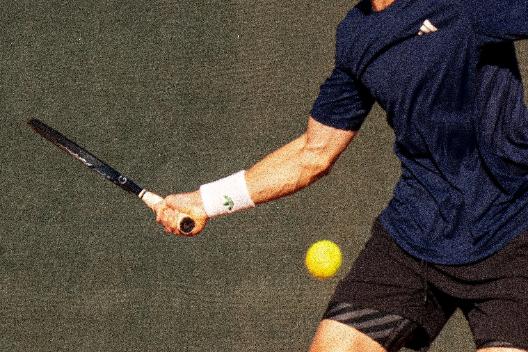 Billy Harris playing tennis, wearing a navy blue shirt and black shorts, holding a tennis racket, with a yellow tennis ball close to their body on a dark court surface.
