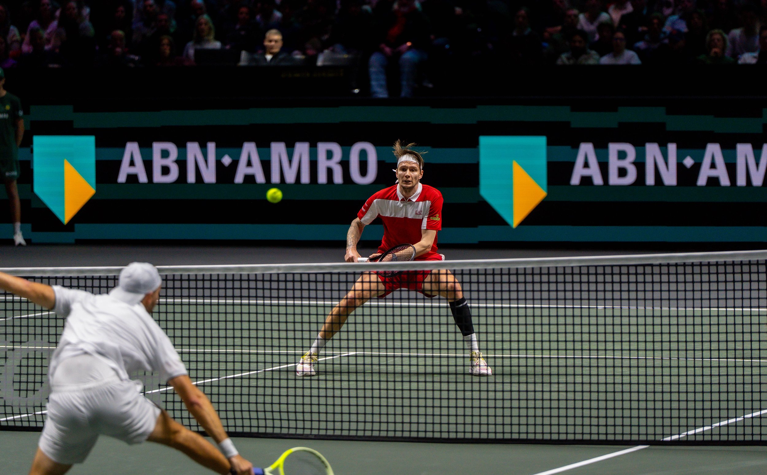 A tennis match with Alexander Bublik, crouching and holding a racket, ready to hit a tennis ball, against a net and a dark background with spectators and digital advertisements.