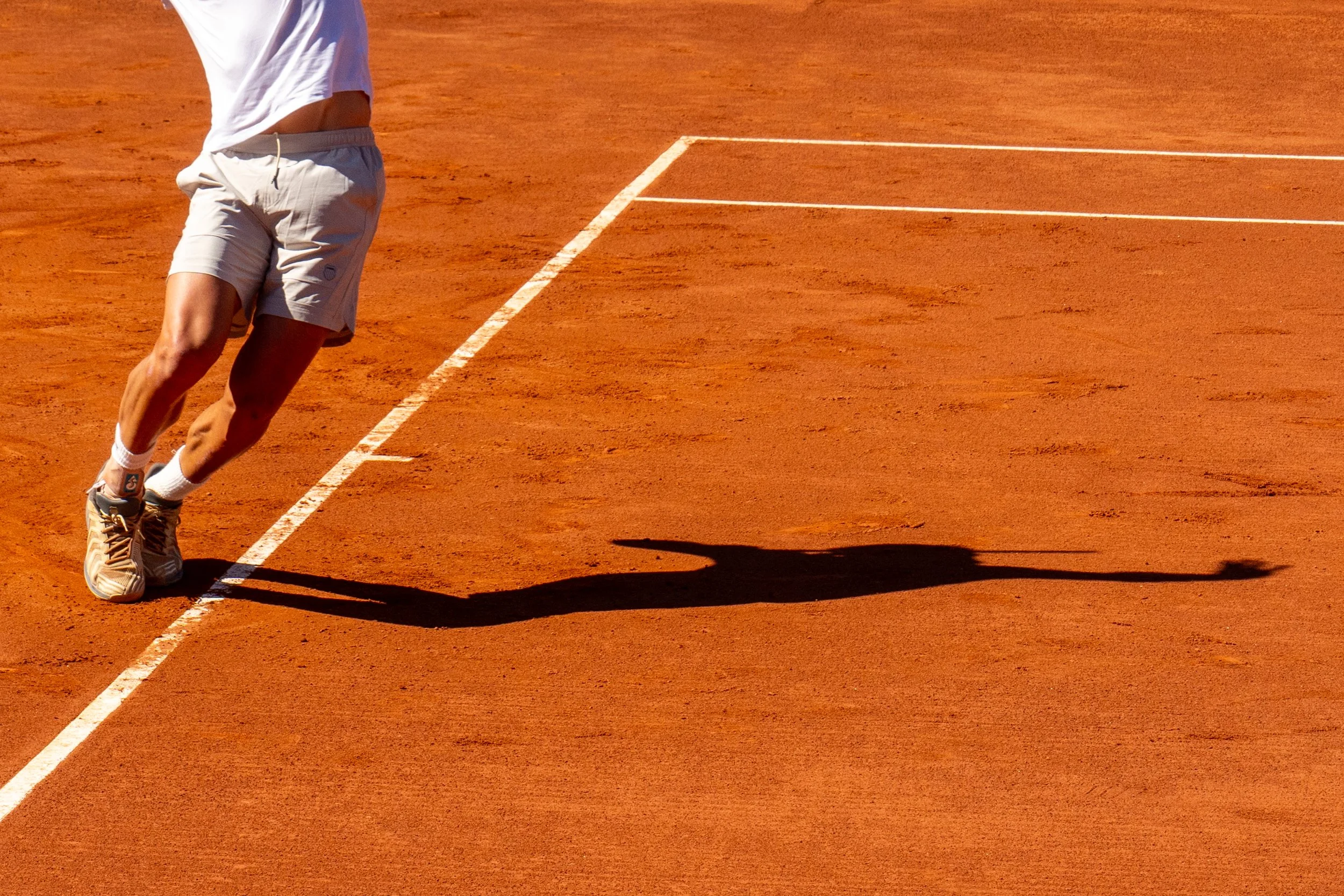 Tomas Etcheverry on a red clay tennis court, with a shadow cast on the ground.