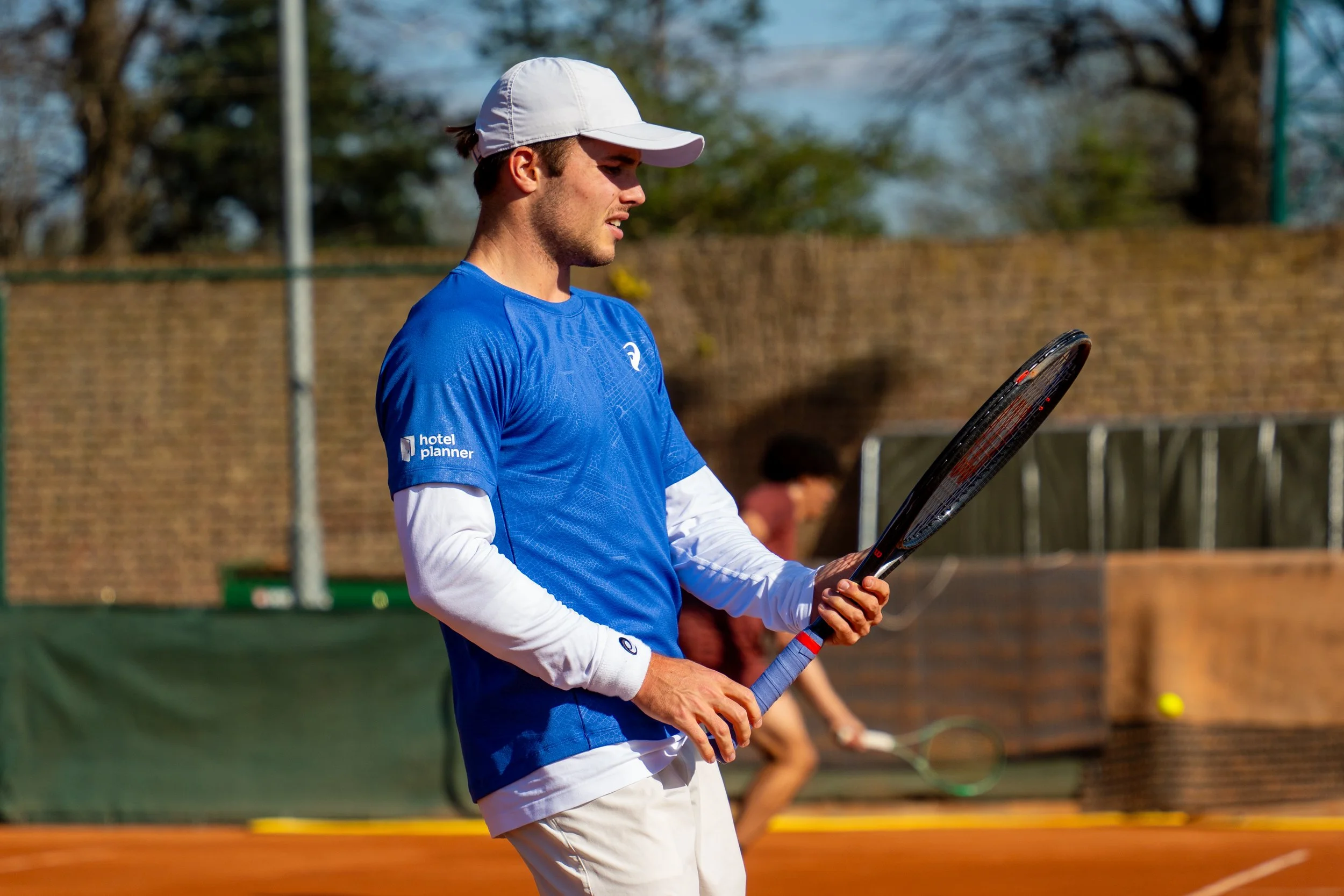 A young man on a tennis court playing tennis, holding a tennis racket, wearing a blue shirt, white sleeves underneath, a white cap, and white pants. There is a woman in the background with a tennis racket.