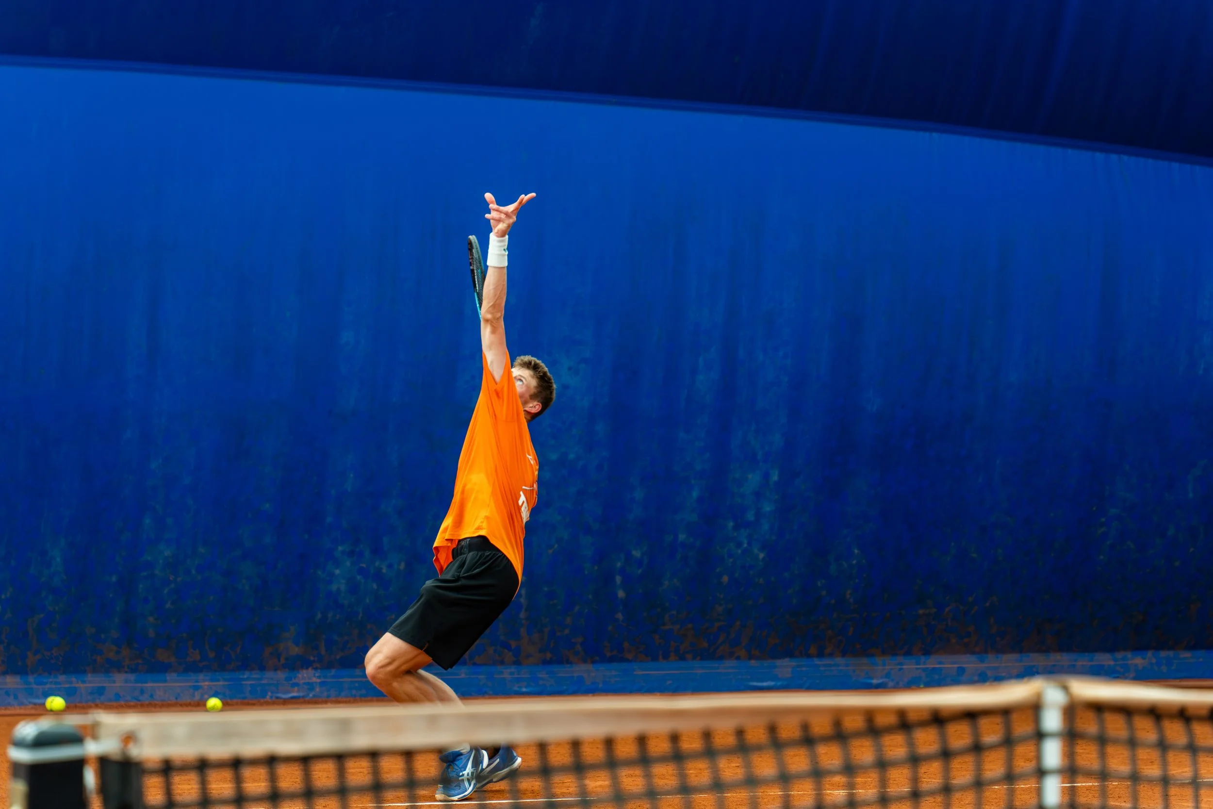 A male tennis player in orange shirt and black shorts about to hit a tennis ball on an indoor clay court with a blue wall background.