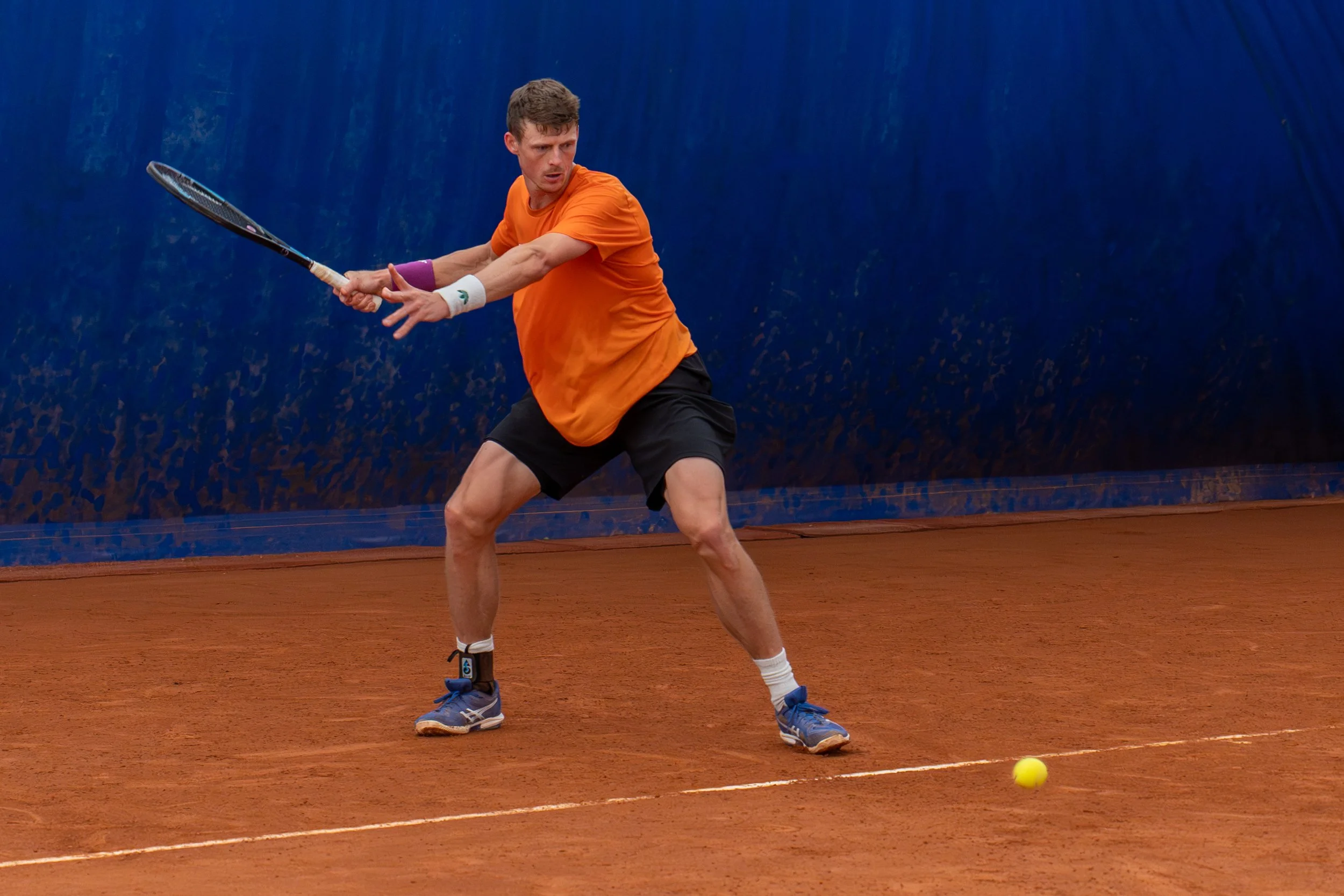 A man playing tennis on a clay court, preparing to hit a low shot with his racket, dressed in an orange shirt, black shorts, and blue shoes.