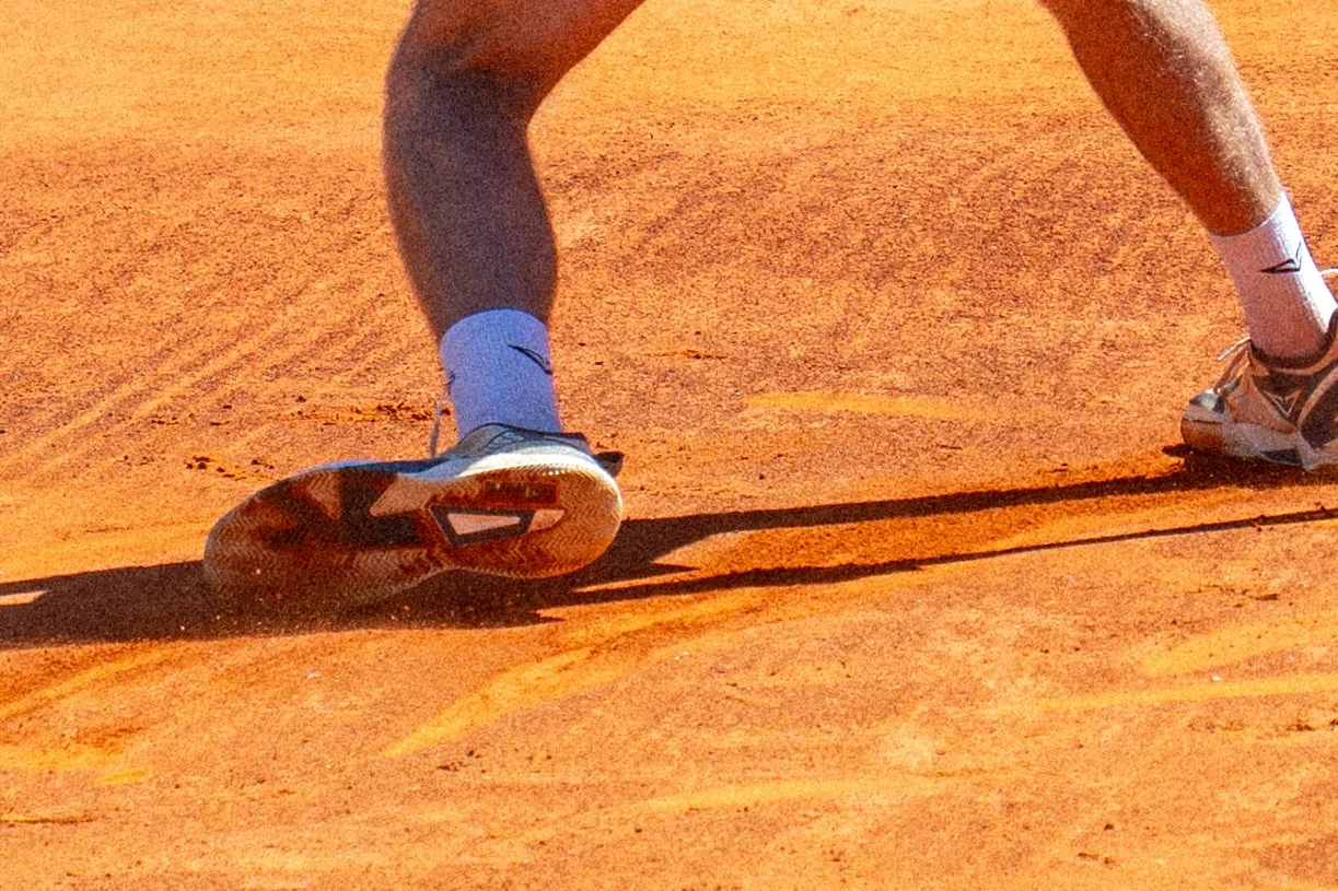 A person playing tennis on a clay court, focusing on their legs and tennis shoes as they slide or move quickly on the court.
