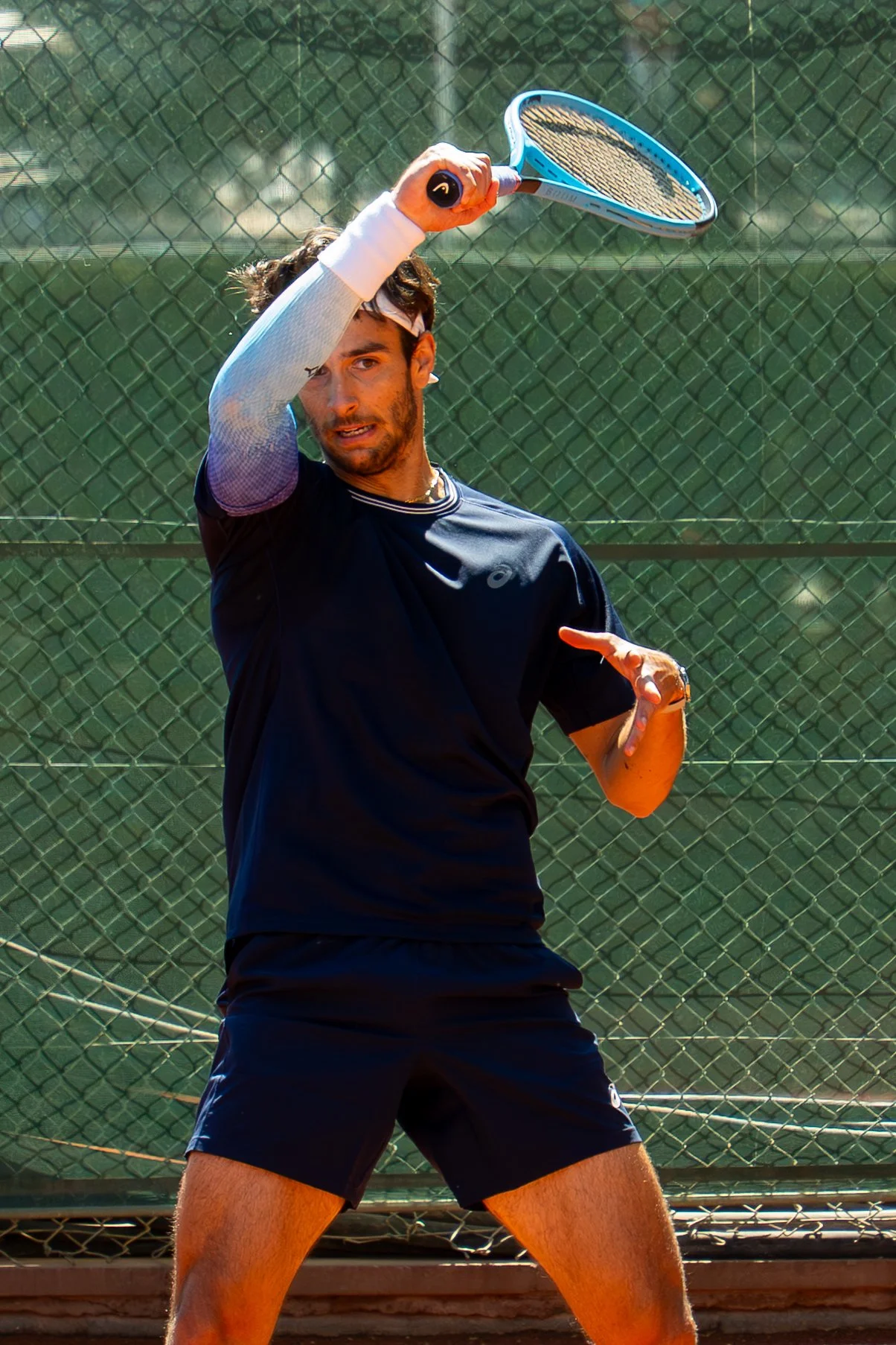 Italian Tennis Player Lorenzo Musetti in black athletic wear and a white wristband, preparing to hit a tennis ball with a blue tennis racquet on an outdoor court with a chain-link fence in the background.