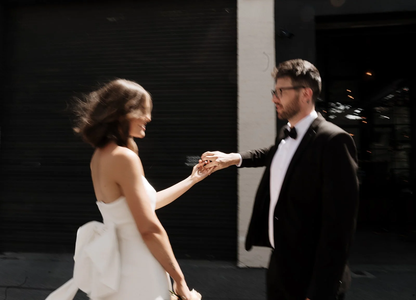 A man in a tuxedo holding hands with a woman in a white strapless dress, they are smiling and appear to be in a joyful moment, outside at night.