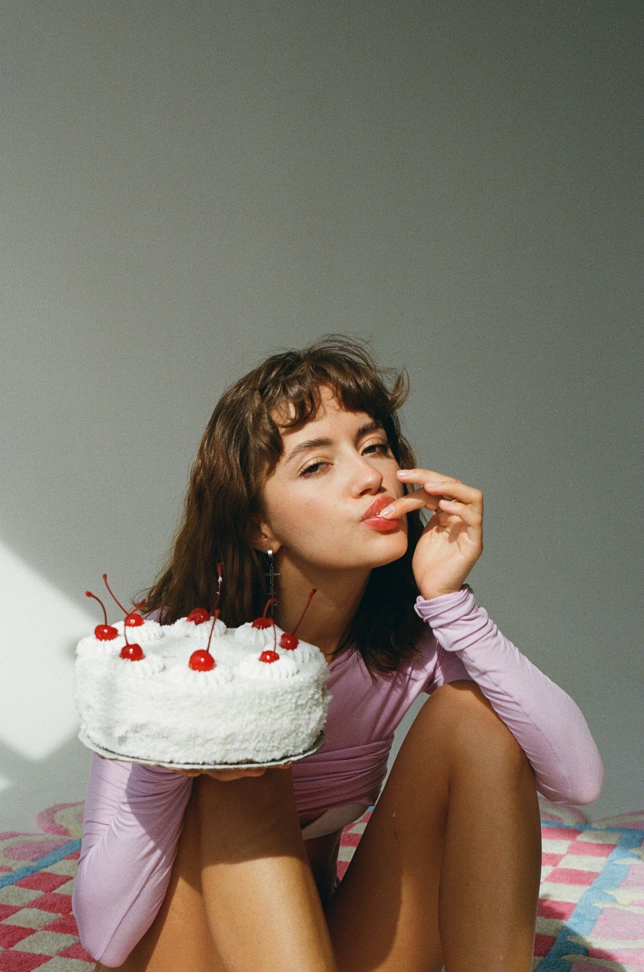 A young woman with short curly hair and light skin sitting on a multicolored checkered mat, holding a white frosted birthday cake with cherries on top, while wearing a lavender long-sleeve shirt and touching her face with her fingers.