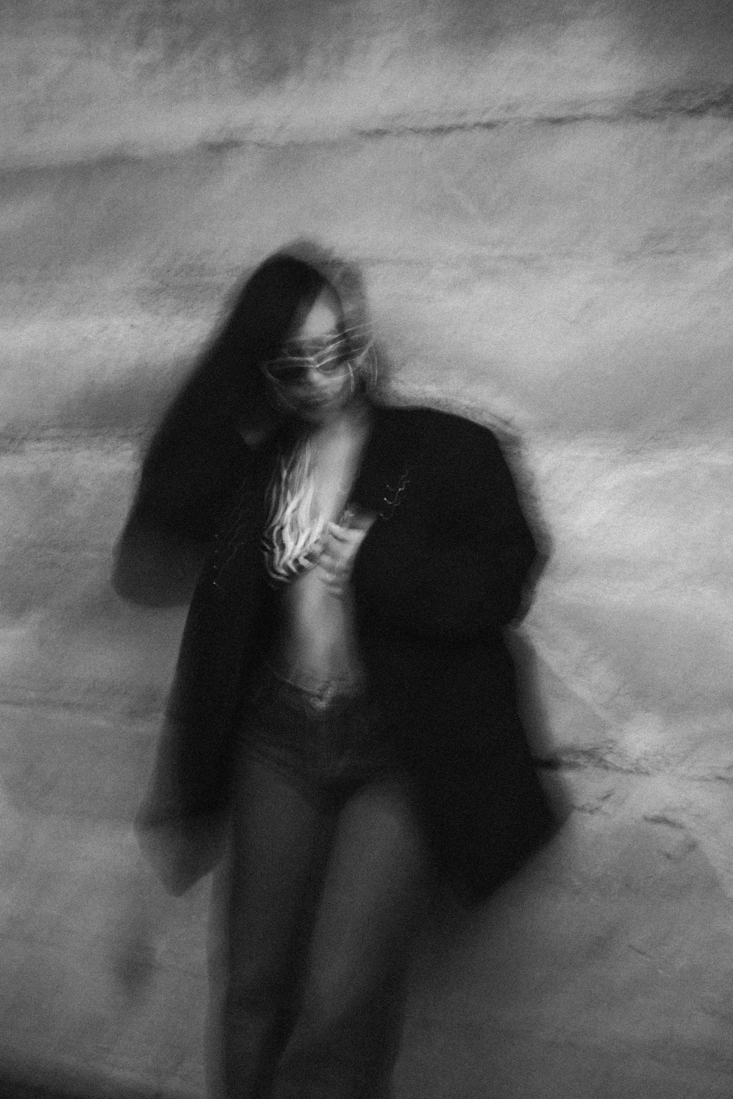 A blurred black and white photo of a person with long hair wearing sunglasses, standing outdoors with a cloudy sky in the background.