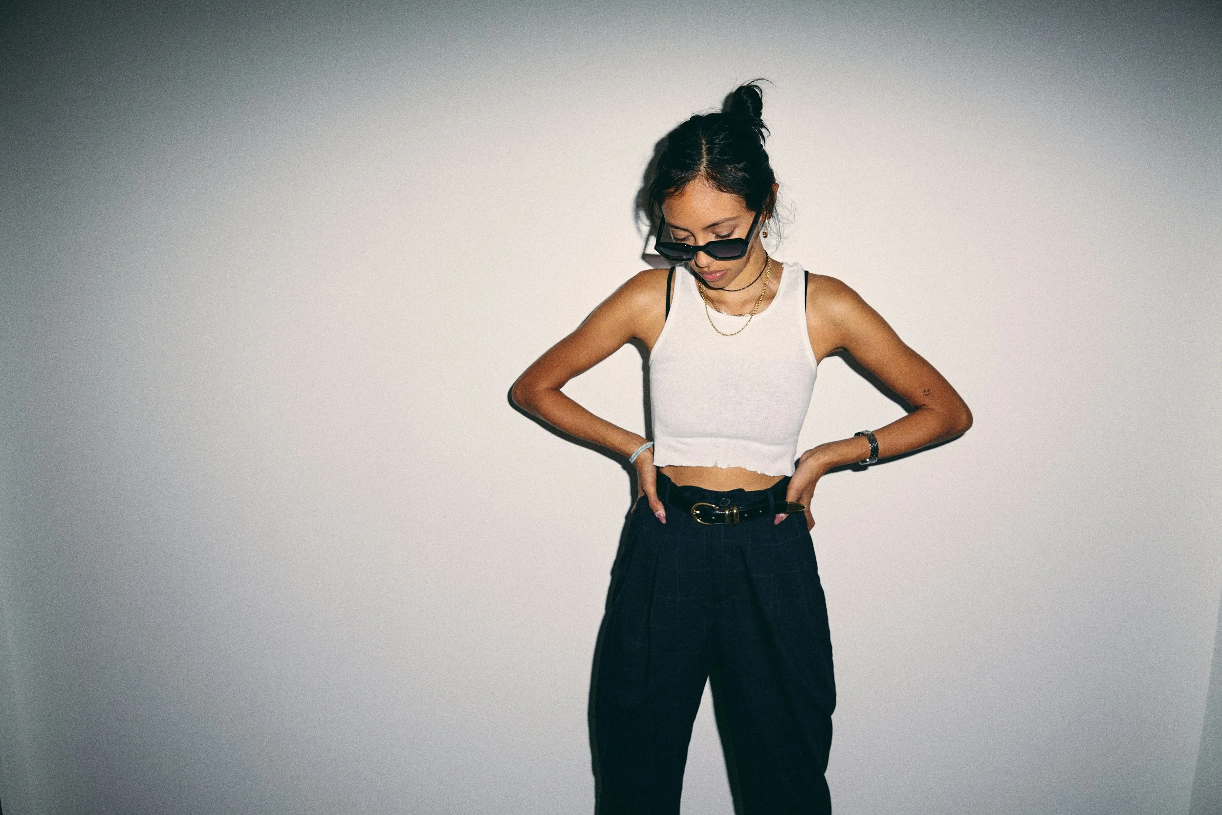 A young woman with dark hair tied up, wearing sunglasses, a white sleeveless crop top, black high-waisted pants, and gold jewelry, looking down with her hands on her hips against a blank wall.