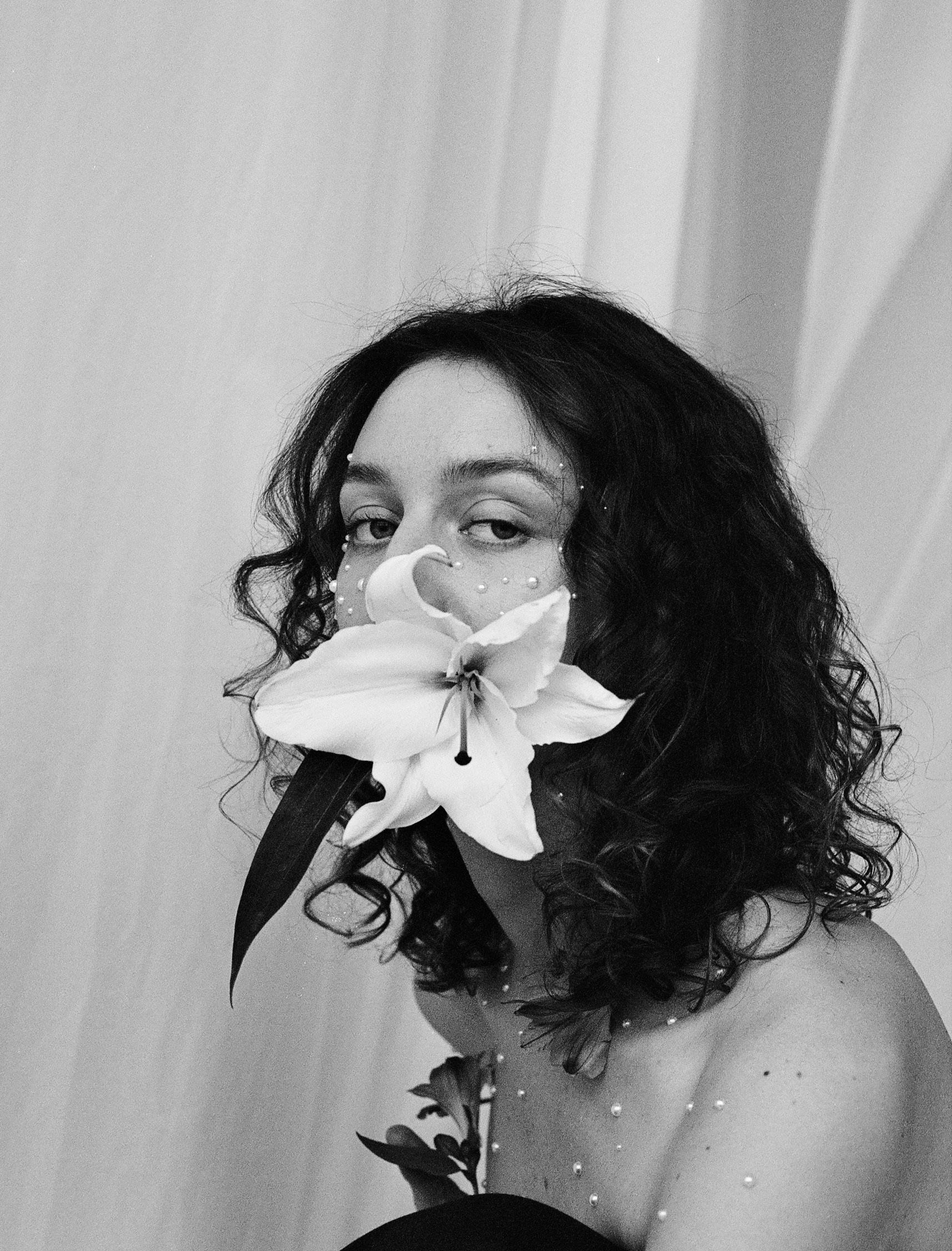A black-and-white photo of a woman with dark curly hair holding a large lily flower close to her face, with pearl-like embellishments on her face and a plain background.