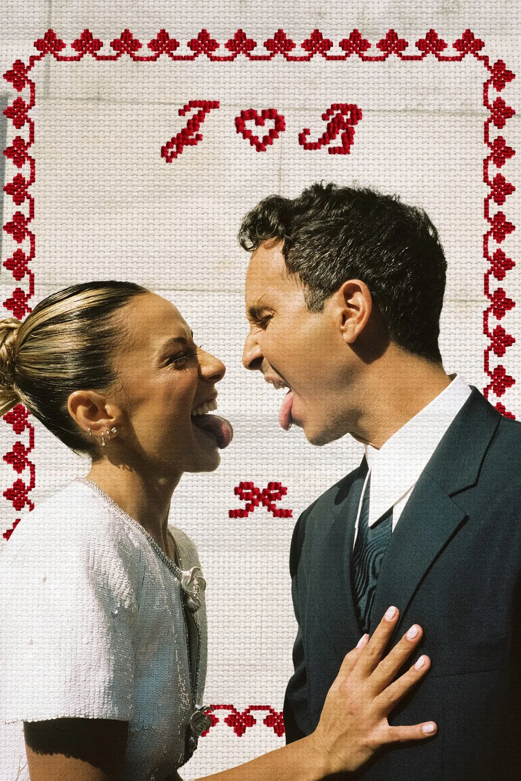 Two people, a woman and a man, are sticking their tongues out and laughing at each other. They are standing close with a decorative cross-stitched wall behind them that reads 'I ♥ J' and features red borders with floral patterns.