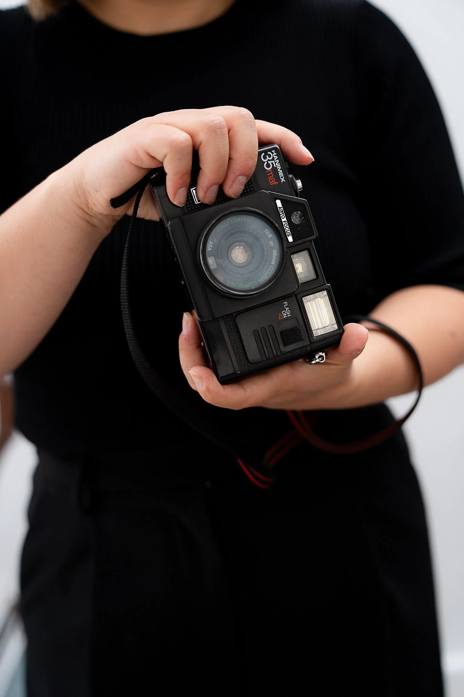 Person holding a vintage black camera with a flash in front of a plain white background, wearing a black long-sleeve shirt.