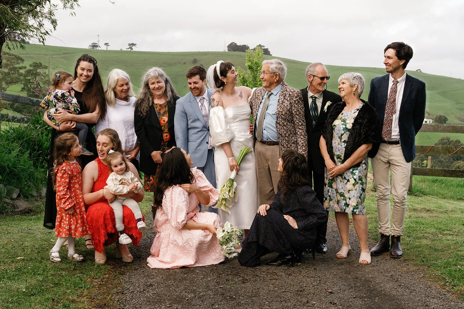 A group of family members gathered outdoors on a cloudy day, with green rolling hills in the background. The bride, in a white dress holding a bouquet of flowers, stands in the center smiling, surrounded by adults and children, all dressed in formal and semi-formal attire, celebrating at a wedding.