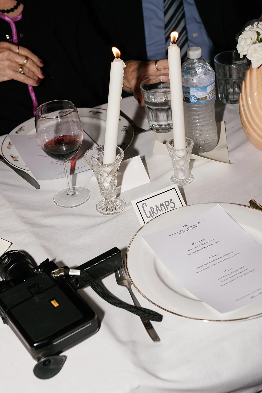 A table setting at a formal event featuring two lit white candles in crystal holders, a glass of red wine, a bottle of water, a menu card, and a card labeled 'GRAmpS'.