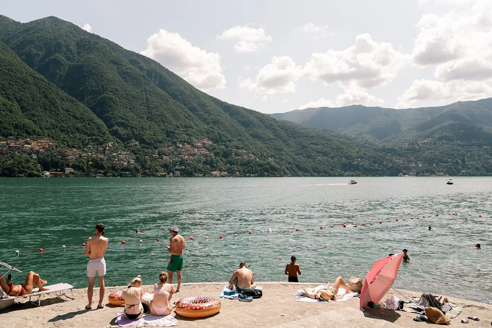 People relaxing and swimming at a lakeside beach with green mountains and cloudy sky in the background.
