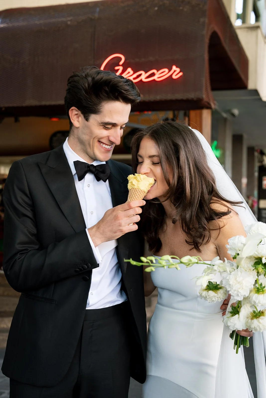 A bride and groom, dressed in wedding attire, sharing a moment with the groom giving the bride a bite of ice cream cone in a restaurant setting.