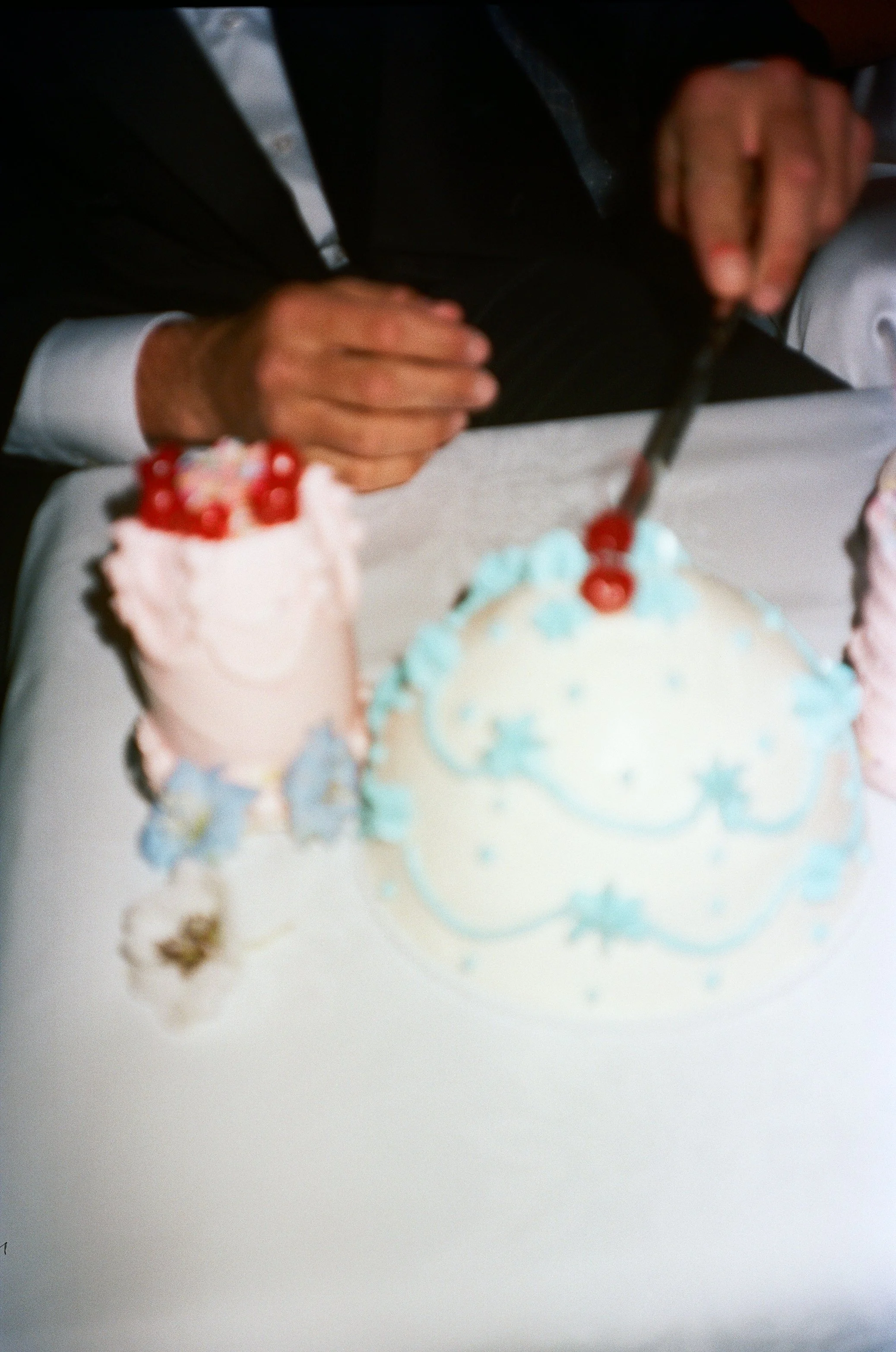 Person in formal attire cutting a birthday cake with white and blue frosting and cherries on top.