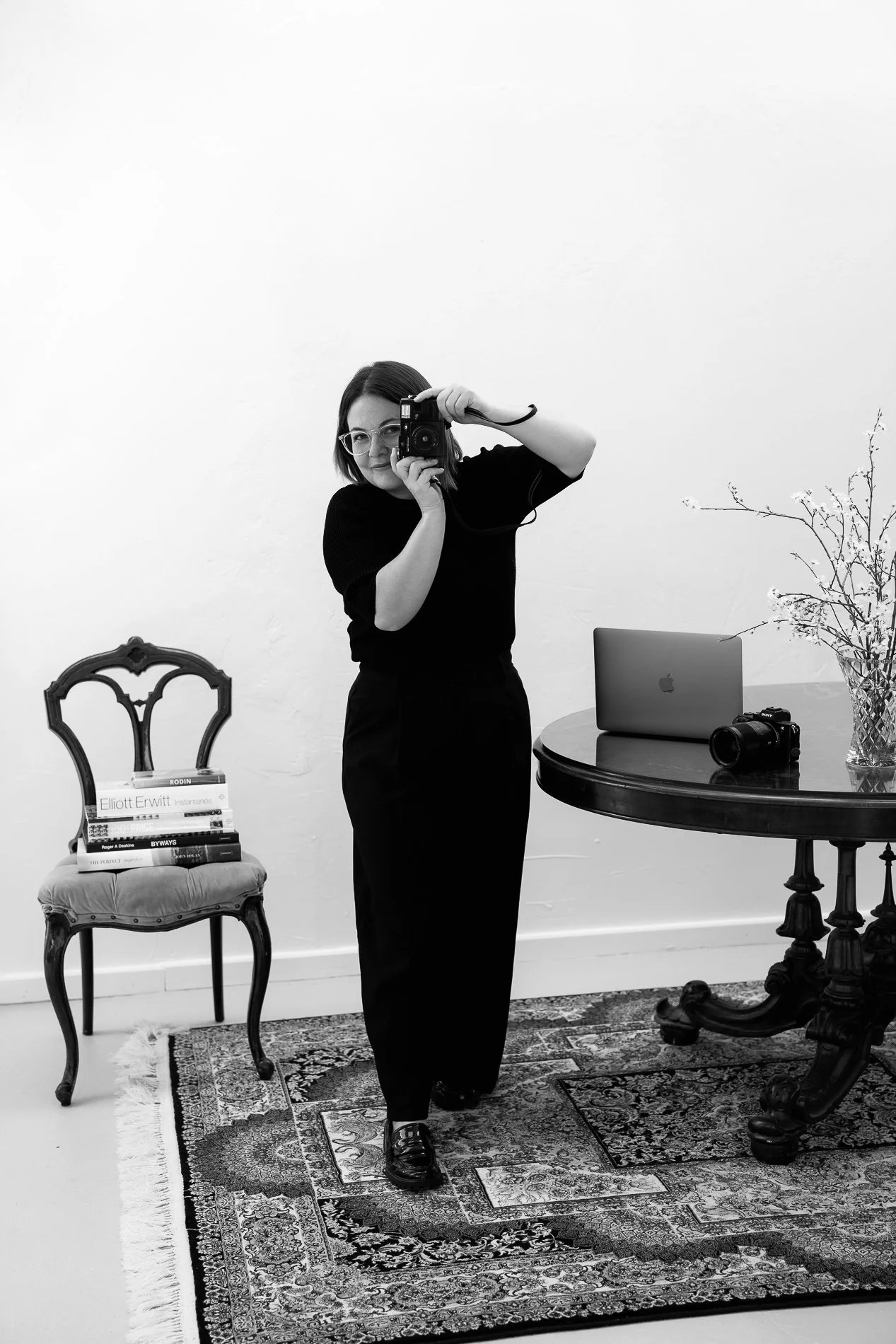 A woman with glasses in black and white, taking a self-portrait with a camera. She stands on a patterned rug, with a small vintage chair with books, a laptop, and a second camera on a round table nearby. The background is plain white with a branch in a vase on the table.