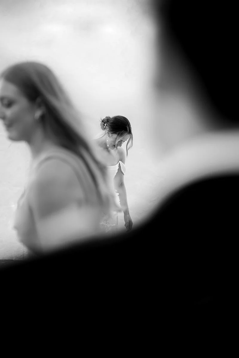 Black-and-white photo of two women, one in the foreground and one in the background, both dressed in sleeveless attire, with the background woman looking down and the foreground woman smiling.