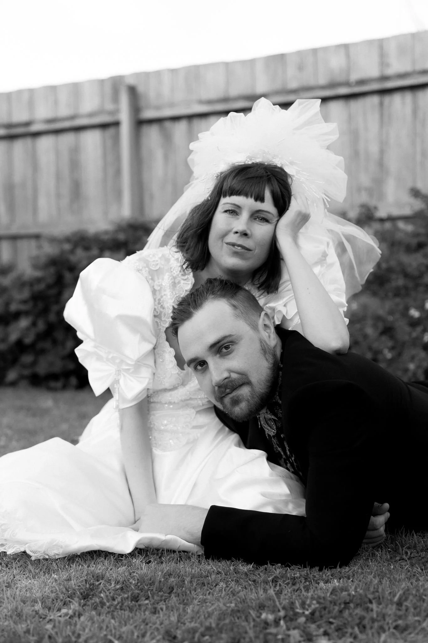 A black and white photo of a woman in a wedding dress and veil, with dark hair and bangs, sitting on the grass next to a man in a suit. The woman is resting her head on her hand and looking at the camera. The man has dark hair, a beard, and is lying on his stomach, propped up on his elbows, looking at the camera. A wooden fence is in the background.