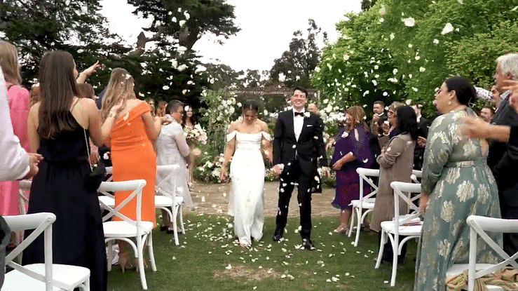 A bride and groom walking arm-in-arm down an outdoor wedding aisle, surrounded by guests throwing flower petals.