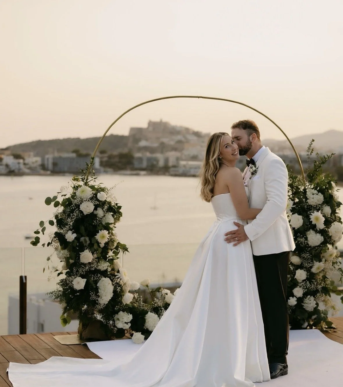 A newlywed couple in wedding attire standing under a decorative arch with white flowers near a body of water during sunset.