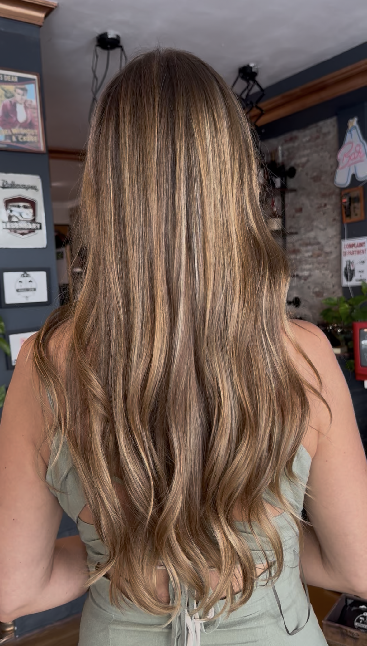 Back view of a woman with long, wavy, light brown hair with highlights, wearing a sleeveless green top, in an indoor setting with a brick wall and decorative signs.