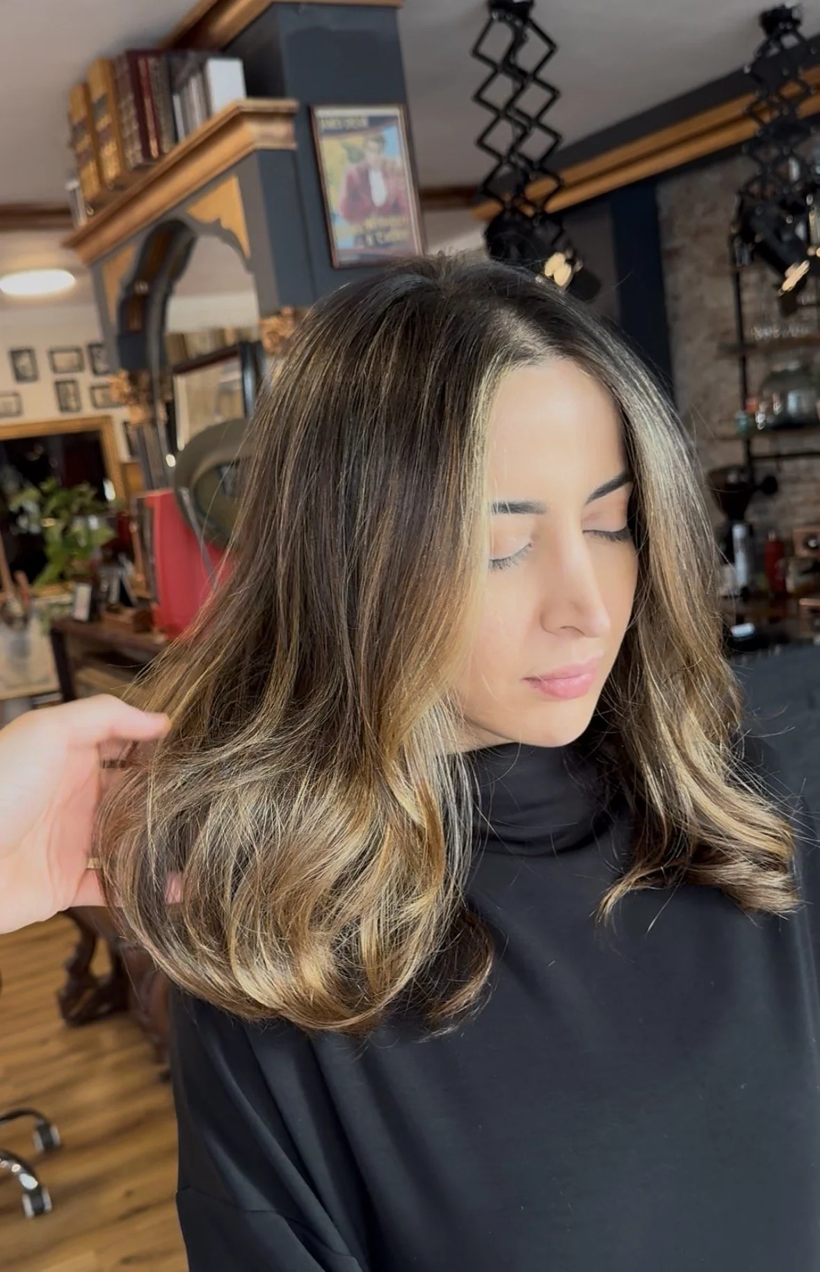 A woman with shoulder-length, wavy hair with highlights, sitting in a salon chair with her eyes closed.