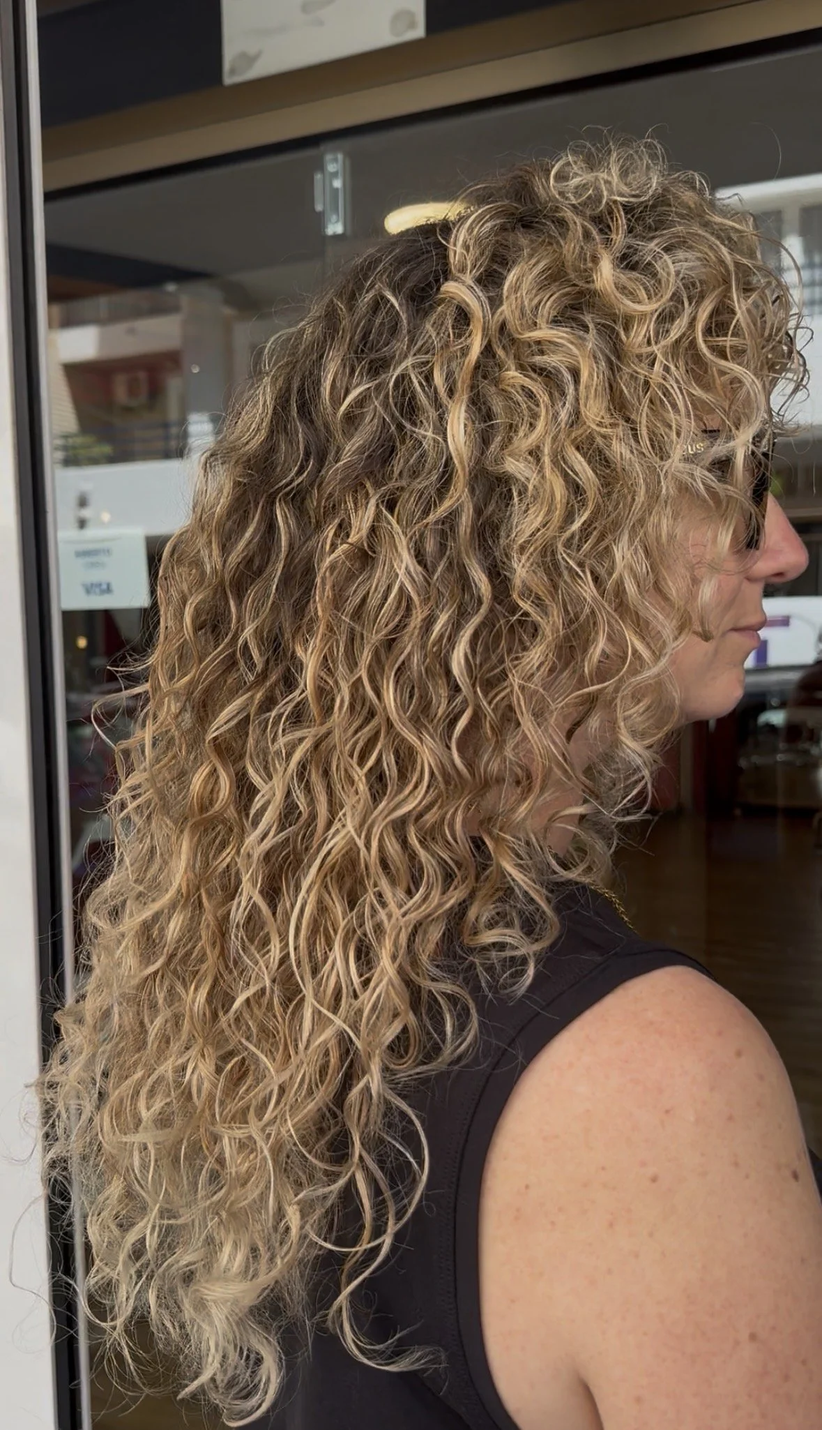 Close-up side view of a woman with curly blonde hair, wearing glasses and a black sleeveless top, standing inside a building near a glass door.