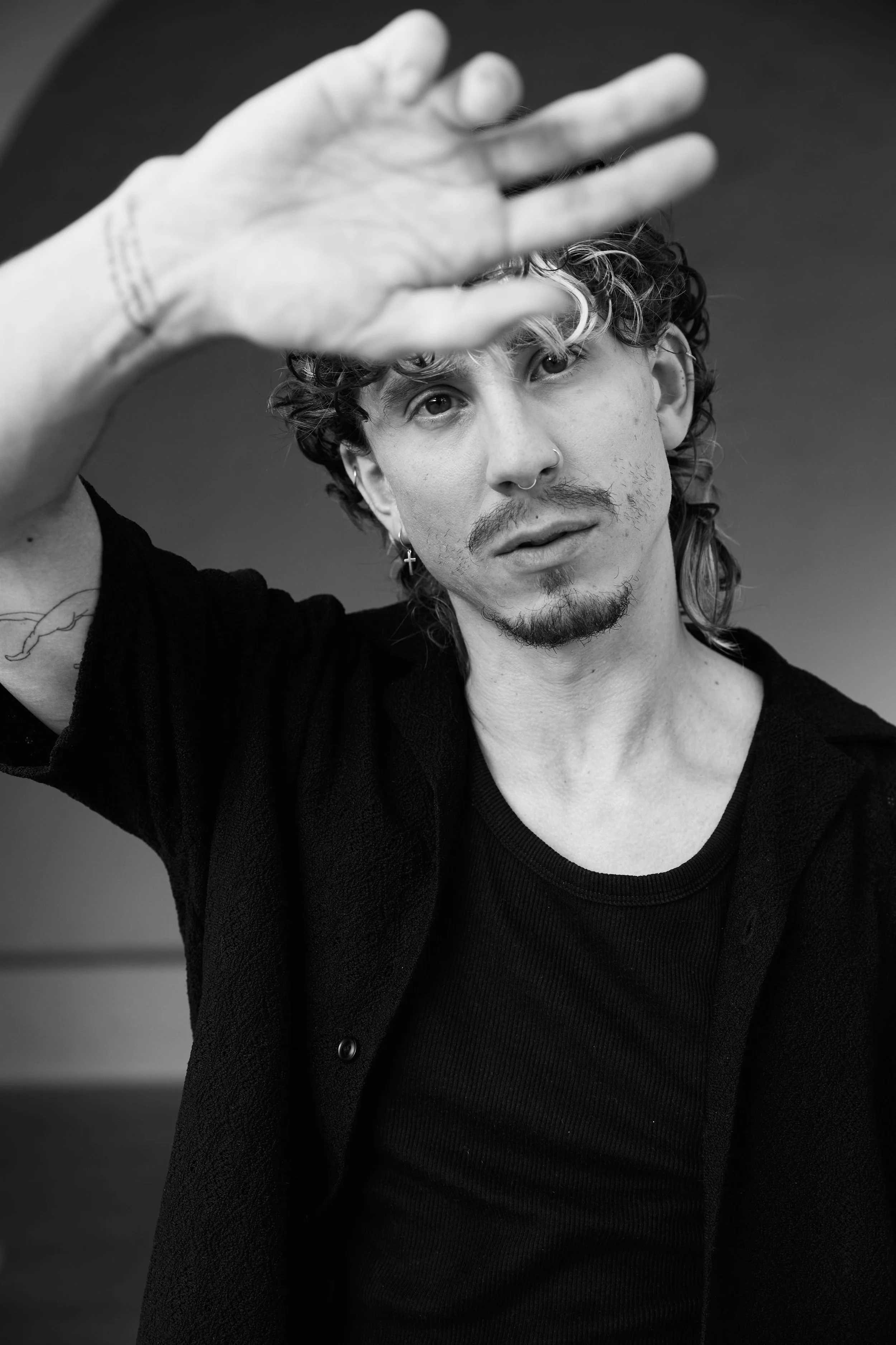 A black-and-white photo of a young man with curly hair, a mustache, and goatee, wearing earrings, raising his hand in front of his face, with a blurred background.