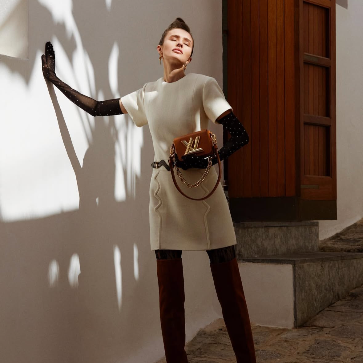 A woman in stylish clothing, wearing a white top and beige skirt, poses with her eyes closed and one arm outstretched, holding a designer handbag with a chain strap, against a wall with shadows and a wooden door in the background.