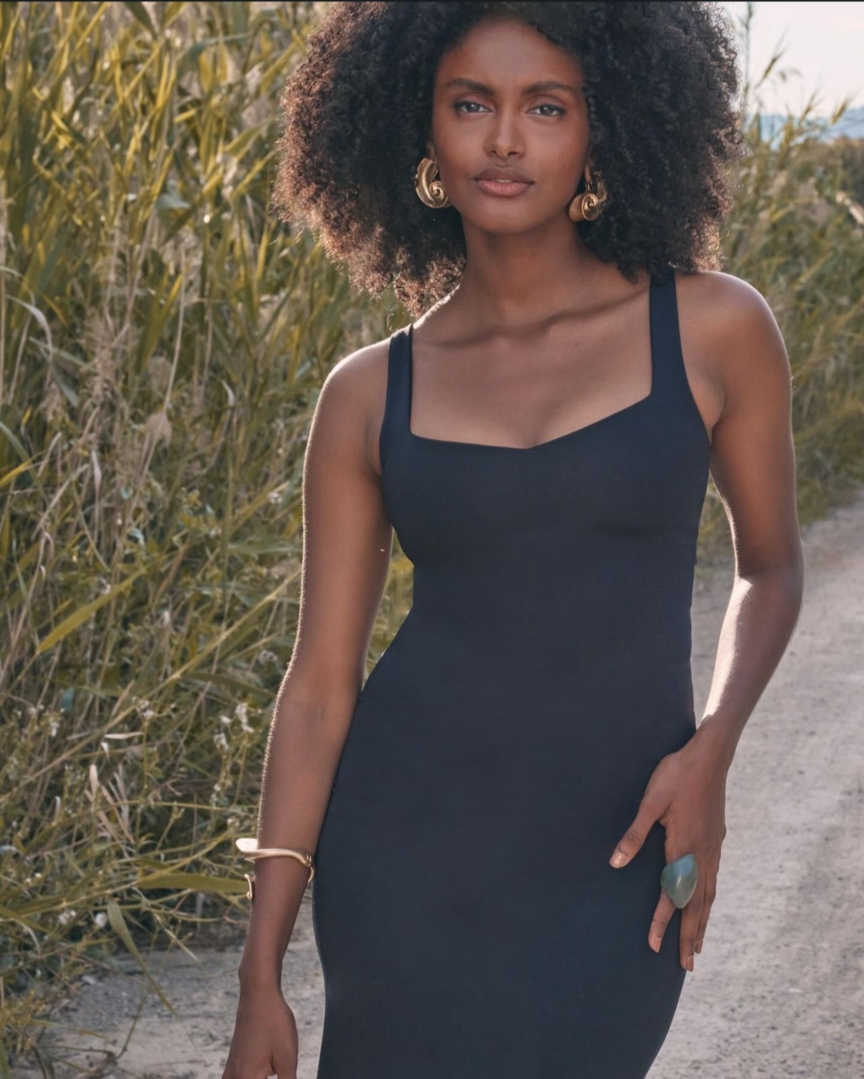 A woman with dark curly hair stands outdoors in front of tall grass, wearing a sleeveless black dress, gold earrings, a gold bracelet, and a large ring, with a confident expression.
