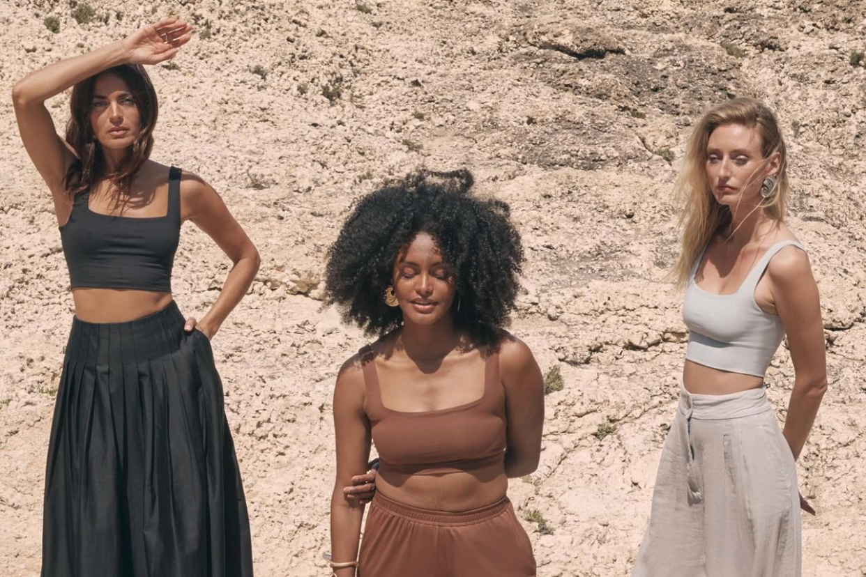 Three women standing outdoors on rocky terrain, with two wearing crop tops and high-waisted bottoms, and one with curly hair and a brown tank top and matching pants.