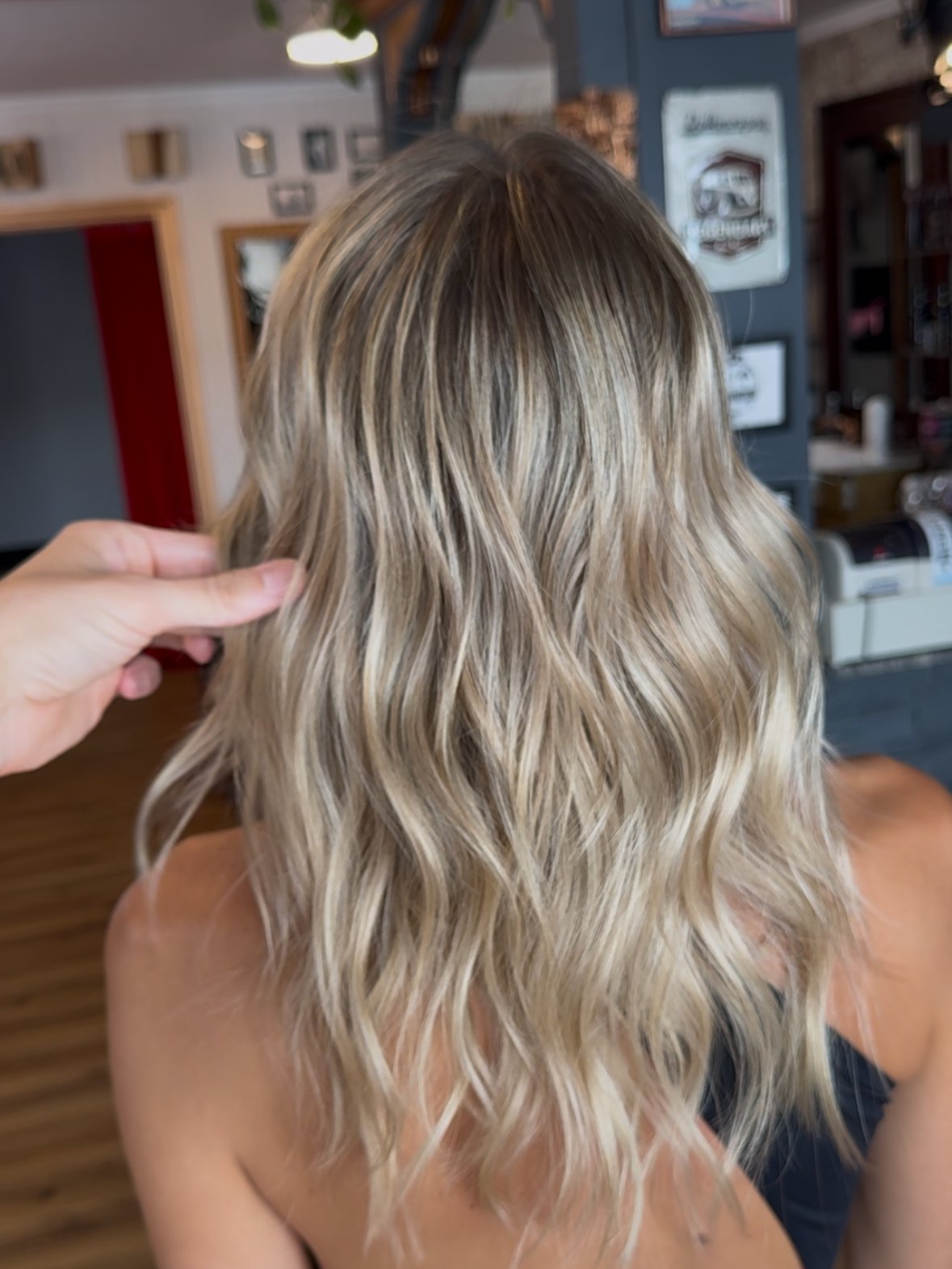 A person with shoulder-length wavy blonde hair sitting in a cozy room, with a hand gently touching the hair.