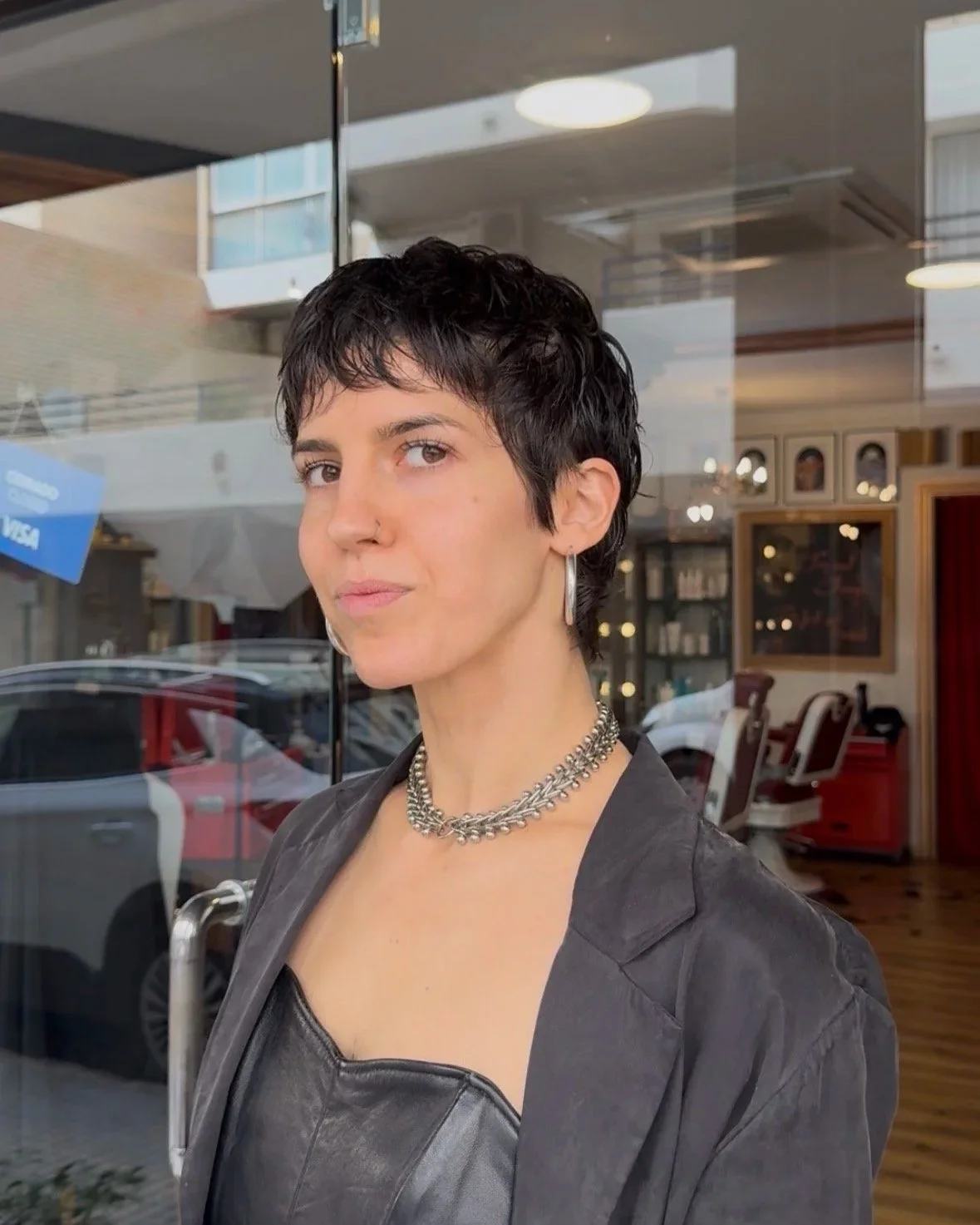 A woman with short, dark hair standing outside a shop window, wearing a black leather top and a chunky silver necklace and earrings.
