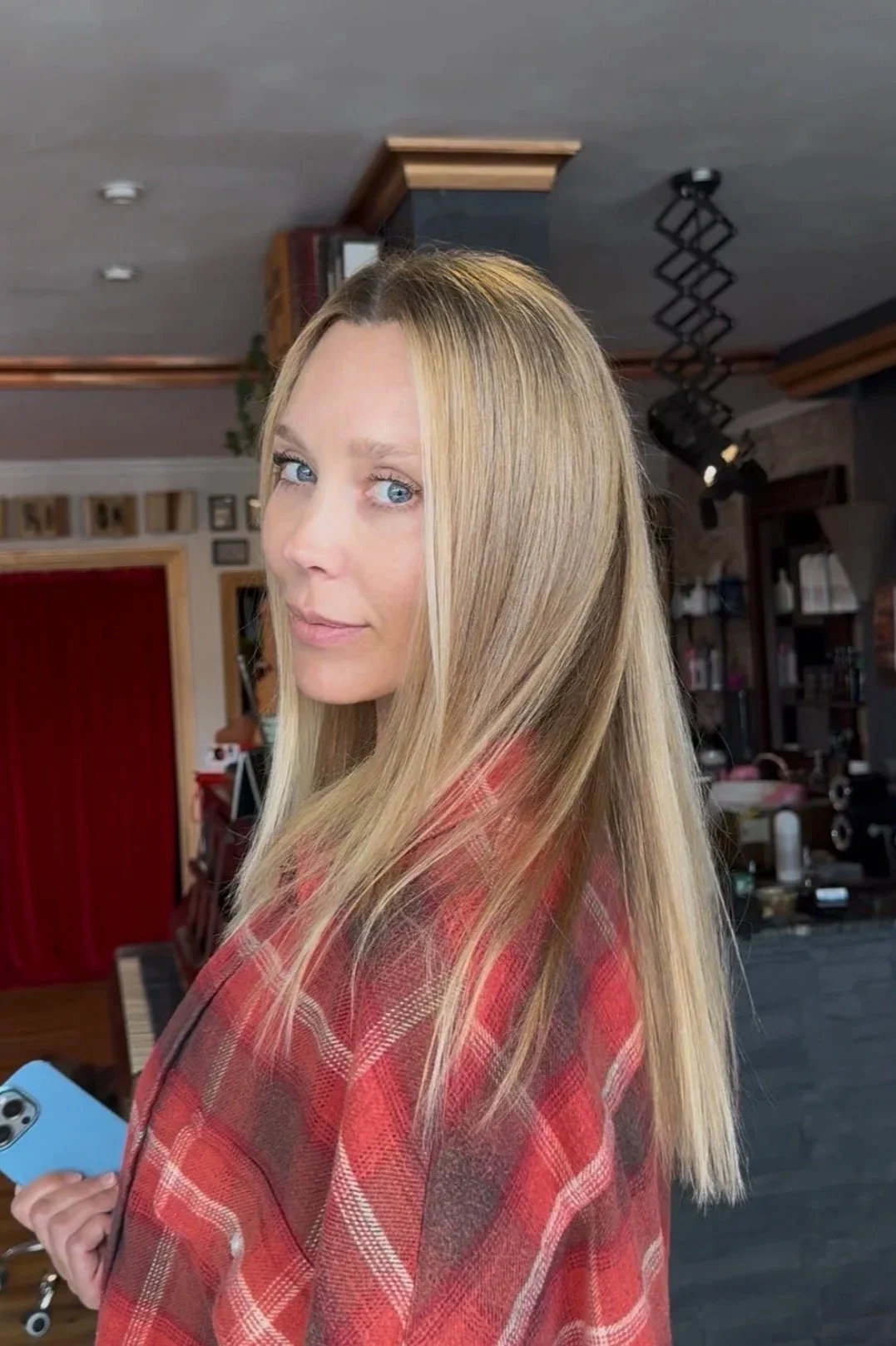 A woman with long, blonde hair and blue eyes holding a blue phone, wearing a red plaid shirt, standing in a room with dark walls and decorative lighting.