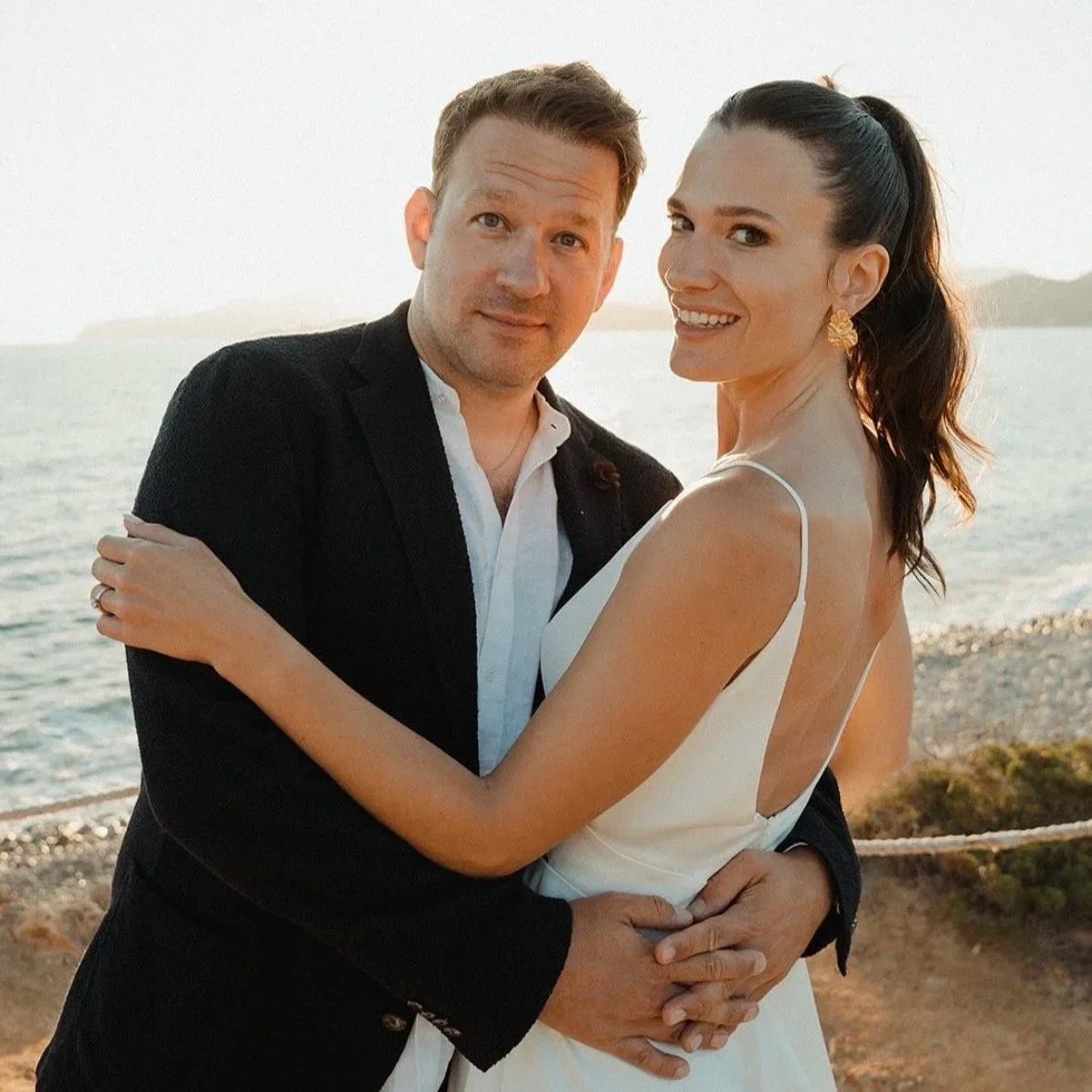 A smiling couple embracing by the water during sunset, the woman in a white dress and the man in a black jacket.
