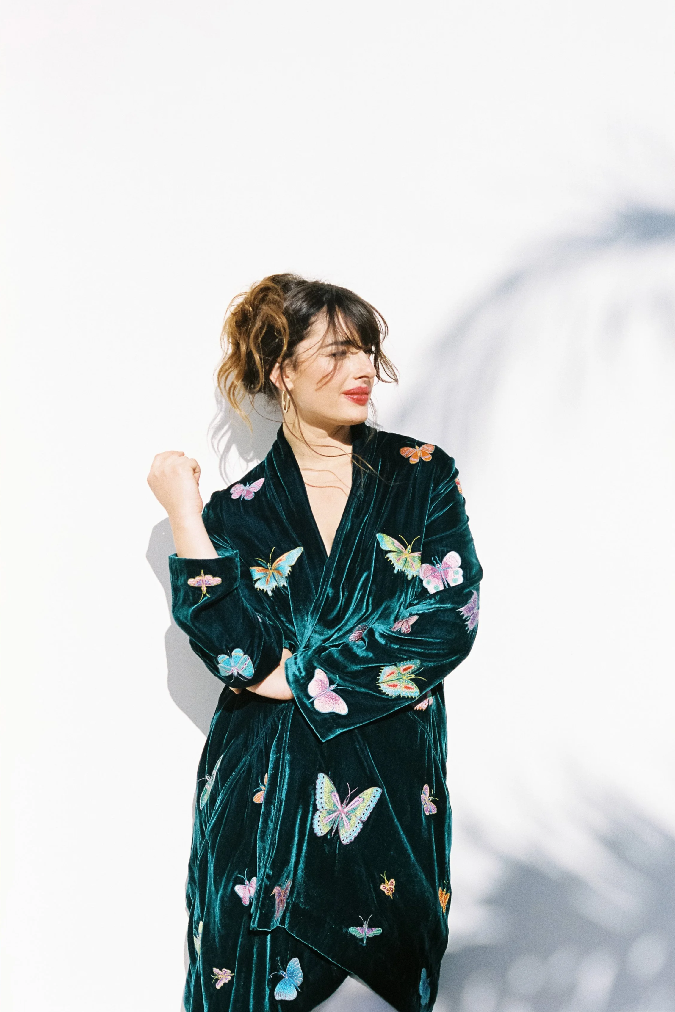 A woman with wavy brown hair dressed in a dark green velvet dress decorated with colorful butterfly embroidery, standing against a plain white background, with her eyes closed and one arm raised in a gesture of celebration or confidence.