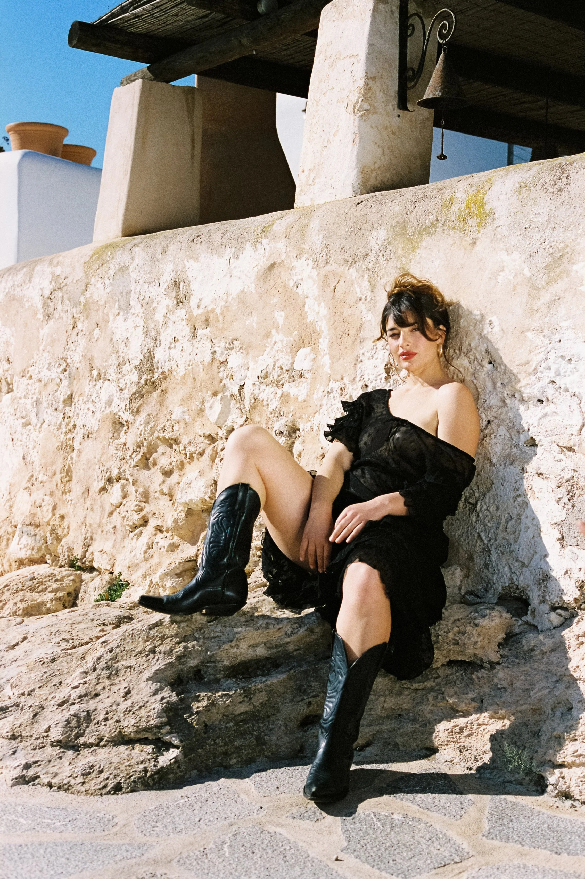 A woman in a black dress and black cowboy boots sitting on rocks against a stone wall outdoors, with a building and blue sky in the background.