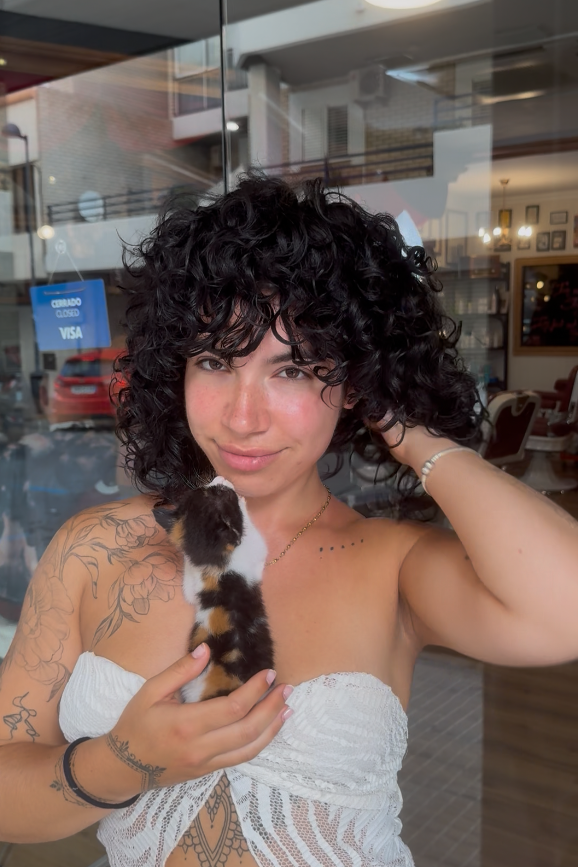 Young woman with curly black hair, tattoos on her arm, wearing a white lace top, holding a calico kitten, standing outside a cafe with a glass window and a sign that says 'Closed' in the background.