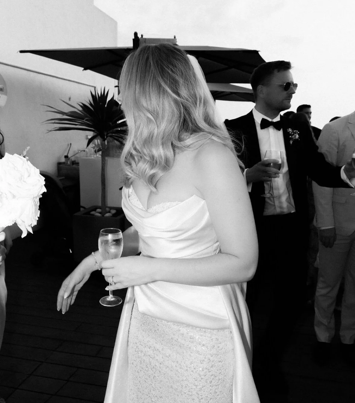 A woman in a wedding dress holding a glass of champagne at a wedding reception, with a man in a tuxedo and sunglasses in the background.