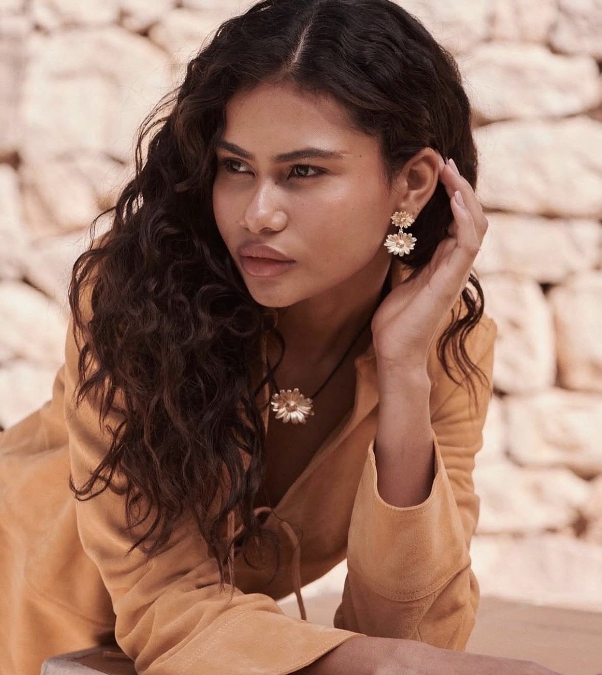 A woman with long curly dark hair wearing flower-shaped earrings and necklace, dressed in a beige or tan top, with a neutral background of a stone wall.