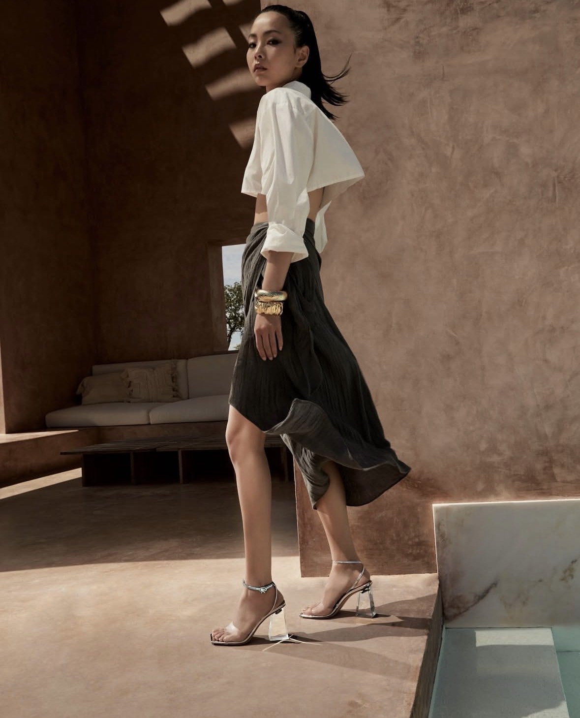 A woman standing indoors, wearing a white blouse, black pleated skirt, and clear high heels, with gold bracelets on her wrist.