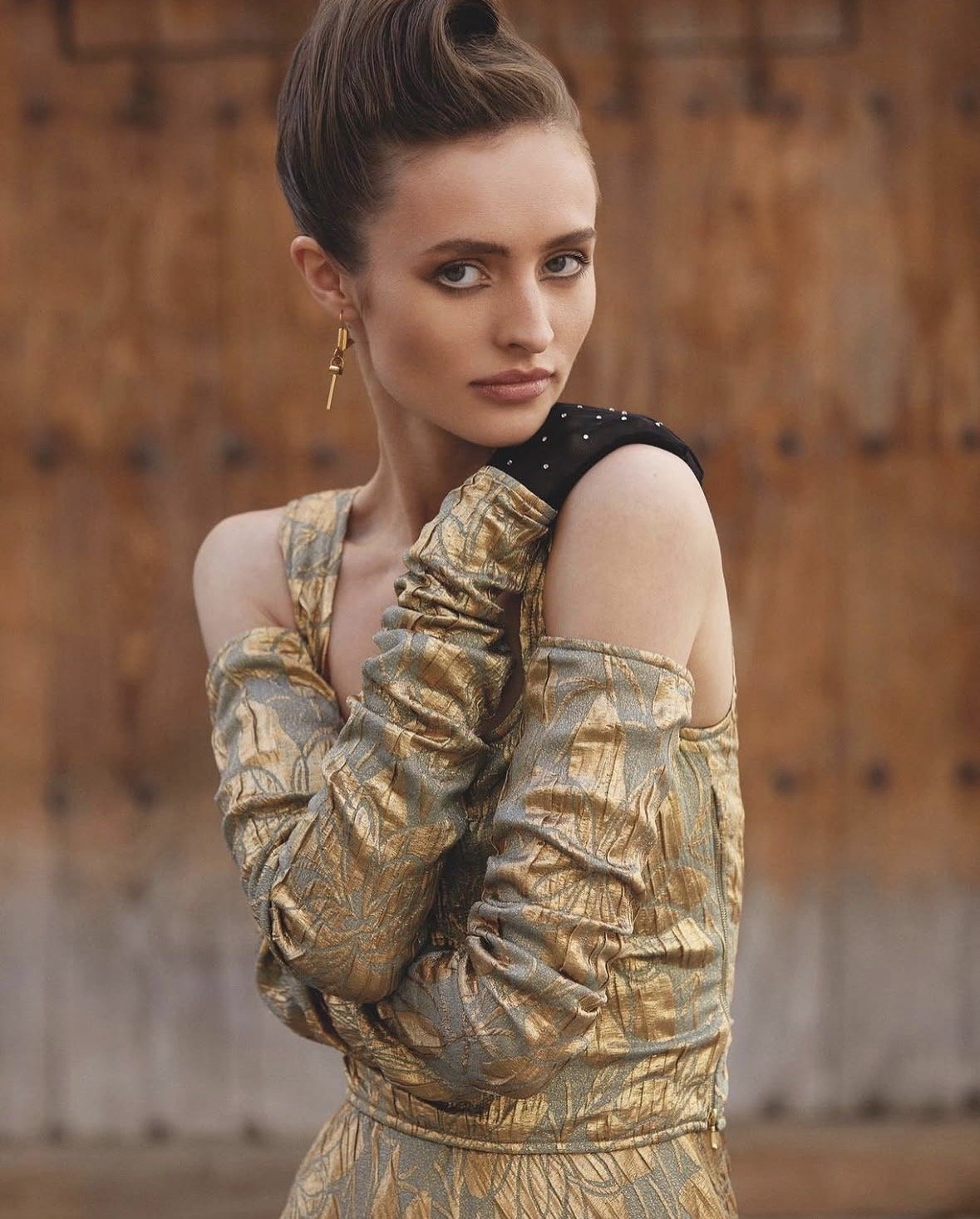 A woman with fair skin, styled brown hair, and makeup, wearing a gold and black off-the-shoulder outfit with ruched sleeves, standing in front of a wooden background.