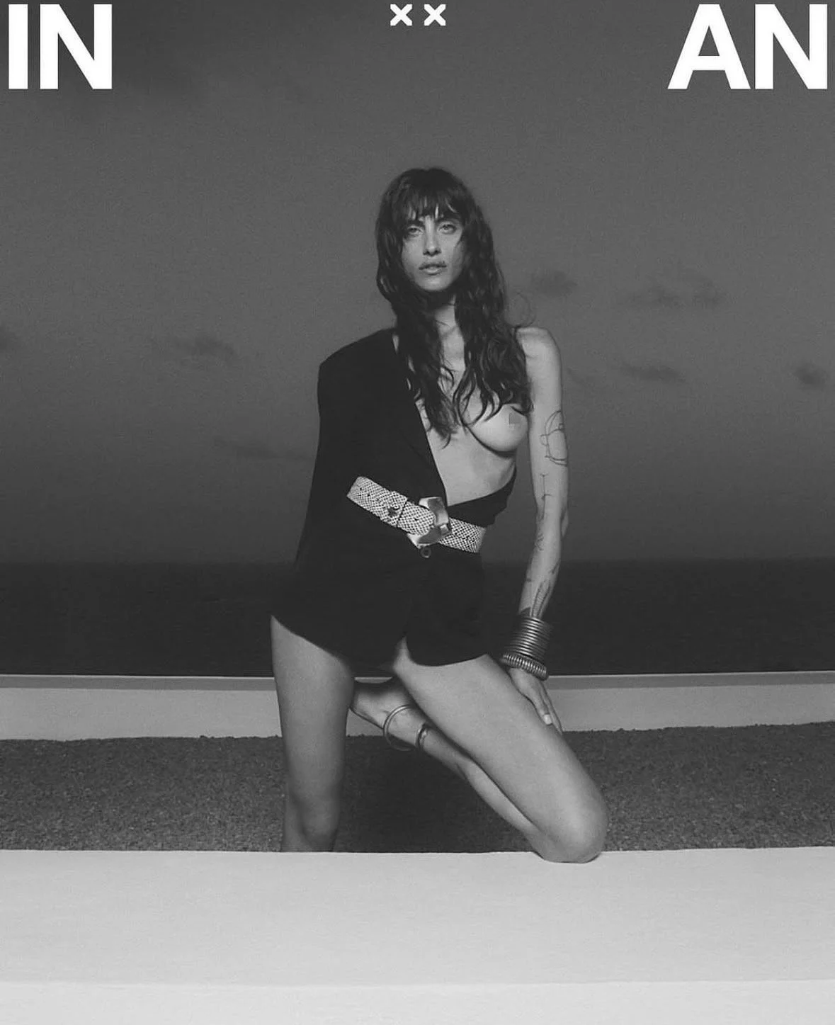 Black and white photo of a woman with tattooed arms and long hair, kneeling outdoors with a cloudy sky background, partially dressed in black clothing.