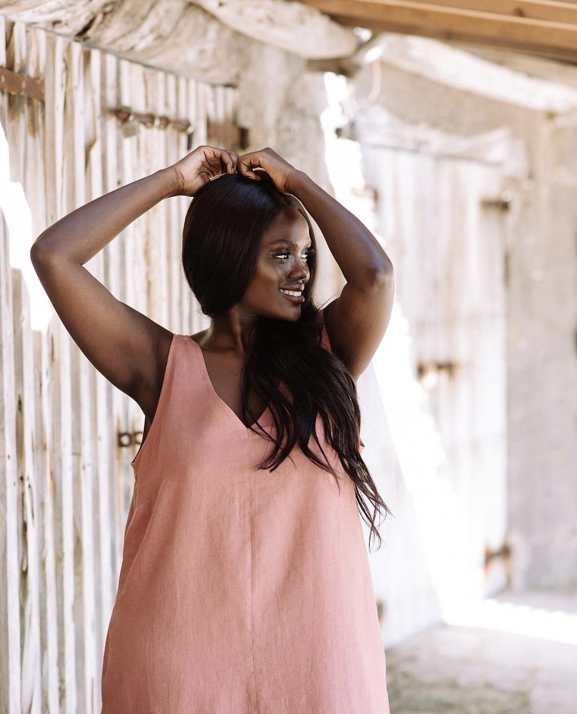 A woman with long dark hair and dark skin wearing a sleeveless pink dress, standing in front of wooden fencing, smiling and touching her hair.