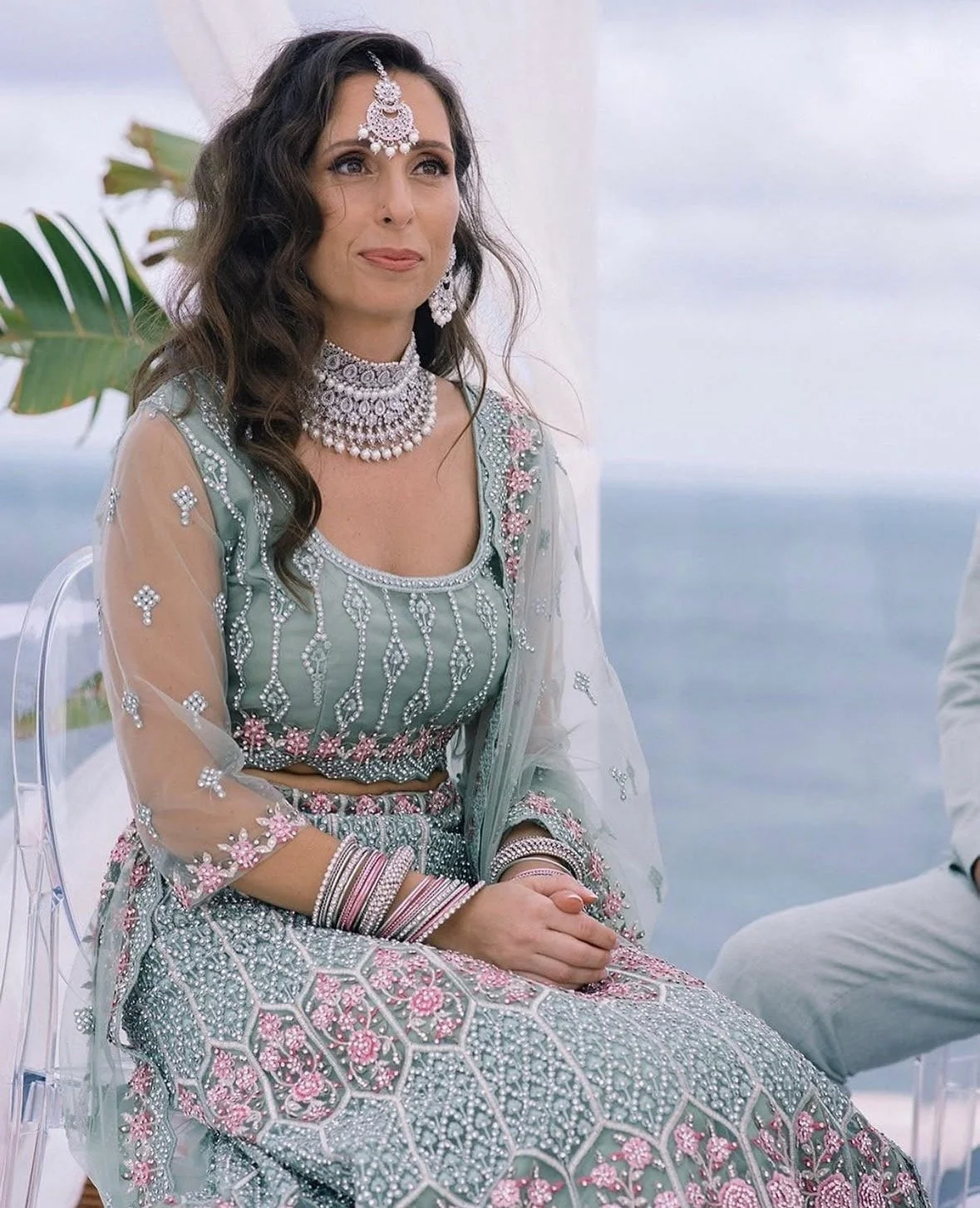 Woman wearing a heavily embellished traditional dress and jewelry, sitting outdoors near the water with a large leaf in the background.