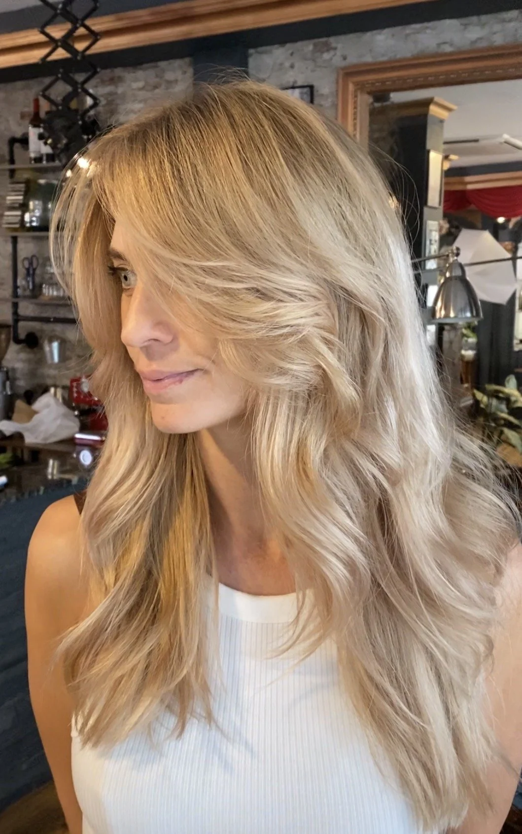 A woman with long, blonde, wavy hair looking to the side, in a room with brick walls and various objects in the background.