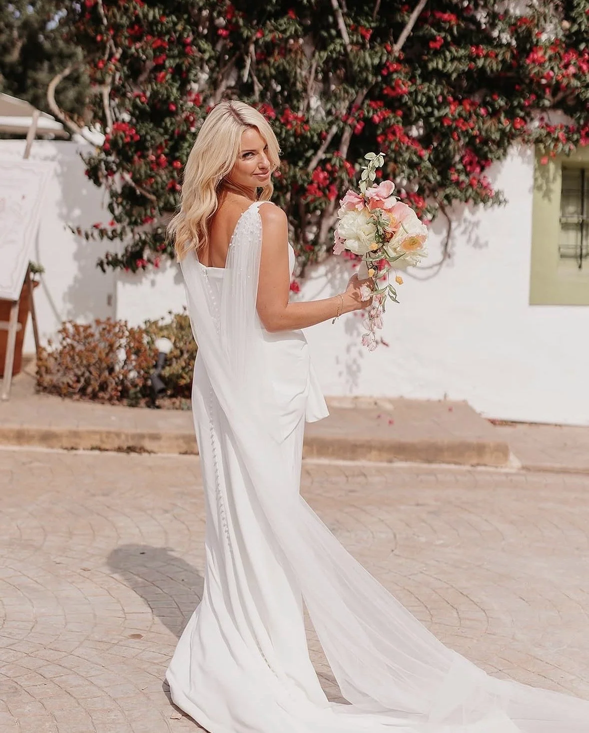 A woman in a white wedding dress holding a bouquet of flowers with a flowering bush in the background.