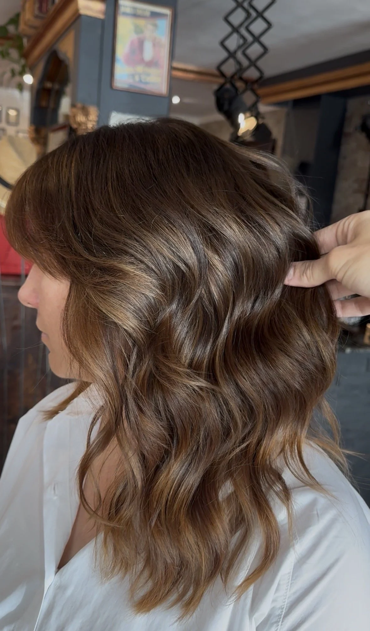 Close-up of a woman's wavy, reddish-brown hair with highlights, held by a hand, in a salon setting.