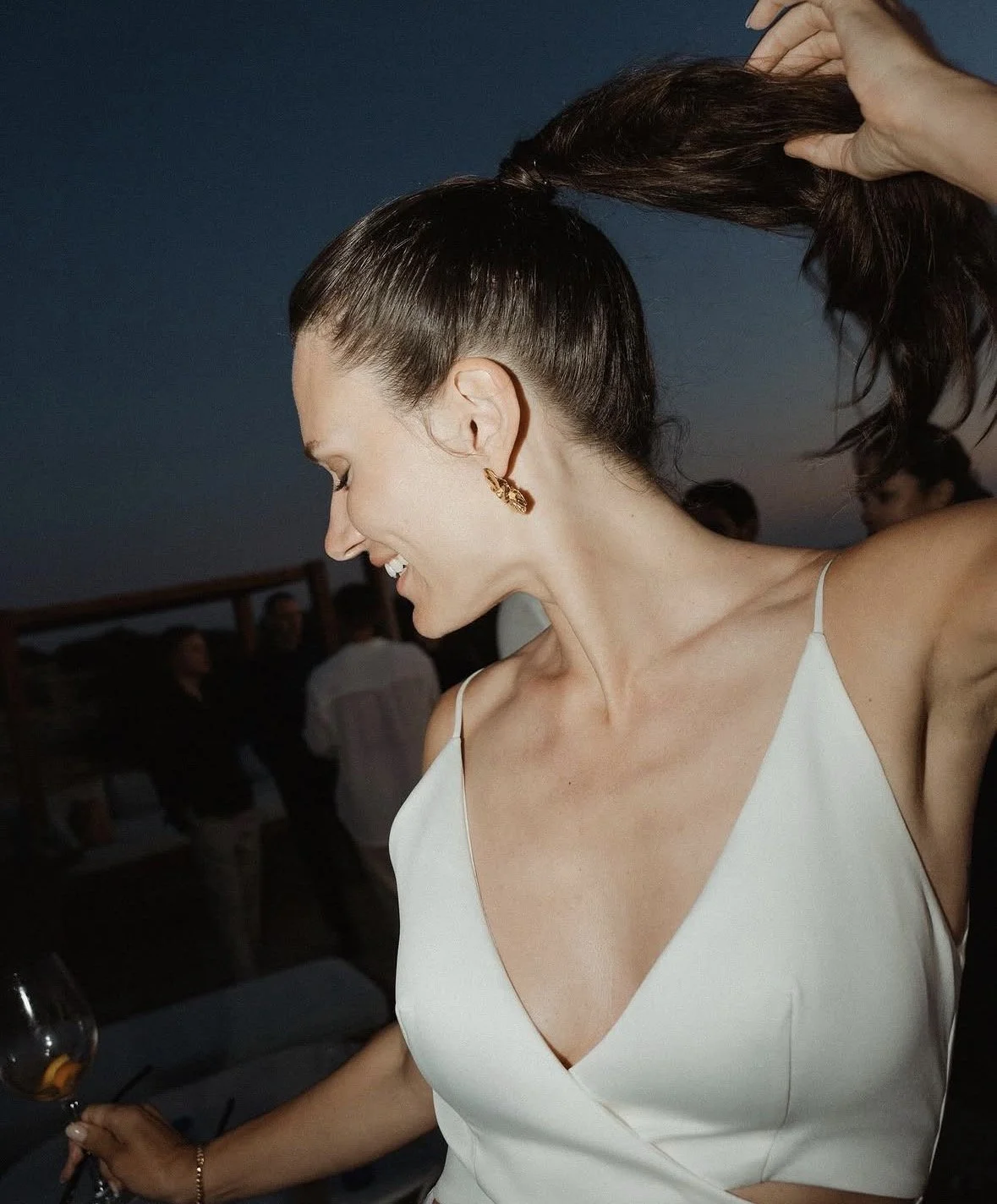 Woman with dark hair in a high ponytail wearing earrings and a white spaghetti strap top, smiling and holding a wine glass at an outdoor gathering during dusk.
